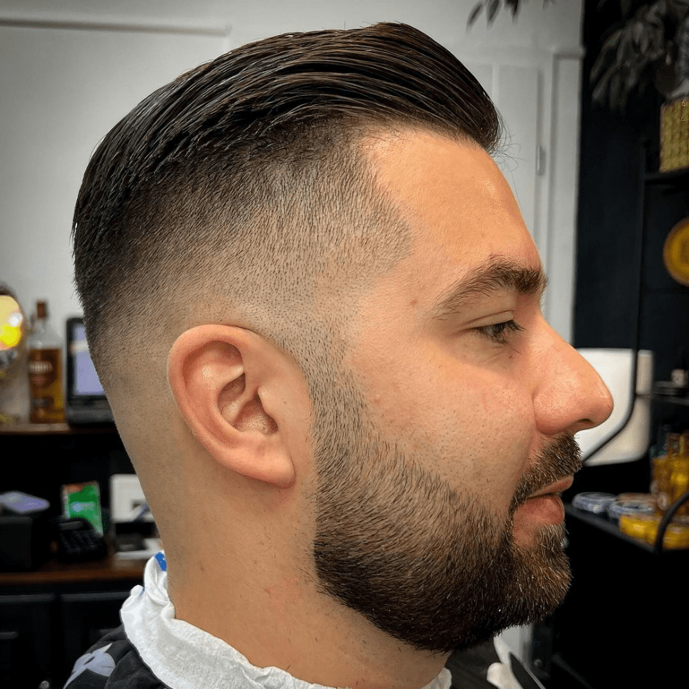 Side profile of a man with a high fade slicked back haircut, featuring sharply faded sides that transition smoothly into the longer, slicked back top. He has a neatly trimmed beard, complementing the modern and clean look. This image highlights a bold and edgy variation of the slicked back hairstyle, perfect for men in 2024 seeking a stylish and contemporary appearance.