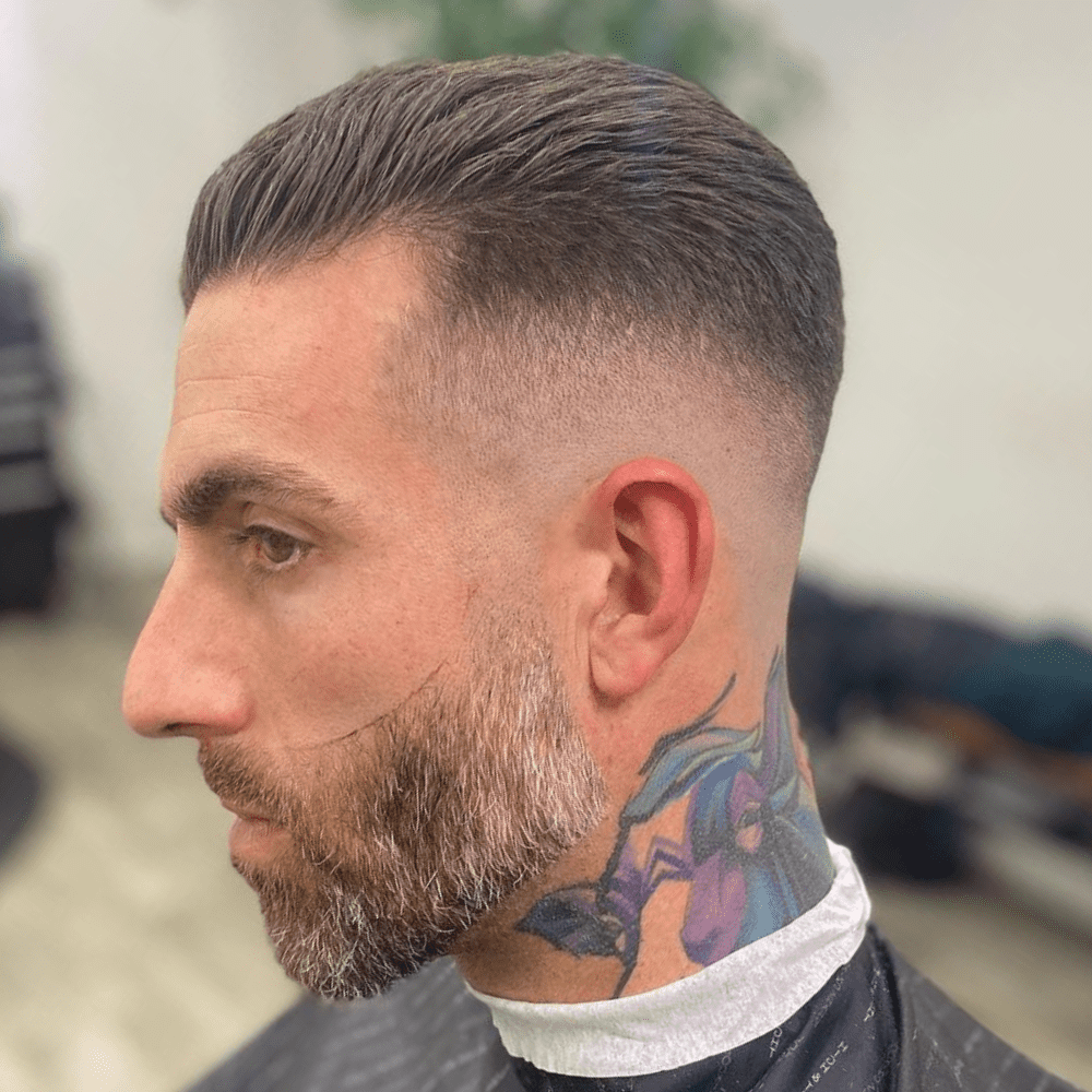 Side profile of a man with a high fade slick back haircut, featuring a smooth fade from the closely shaved sides to the longer hair on top that is slicked back. The style is complemented by a well-groomed beard and a colorful neck tattoo, enhancing the overall polished and edgy look.