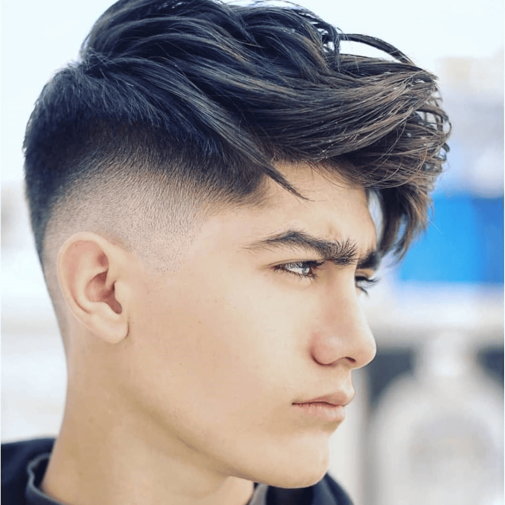 Side profile of a young man with a high fade undercut haircut, featuring a dramatic contrast between the closely shaved sides and the long, textured hair on top. The hair is styled with volume and movement, creating a modern and edgy look.