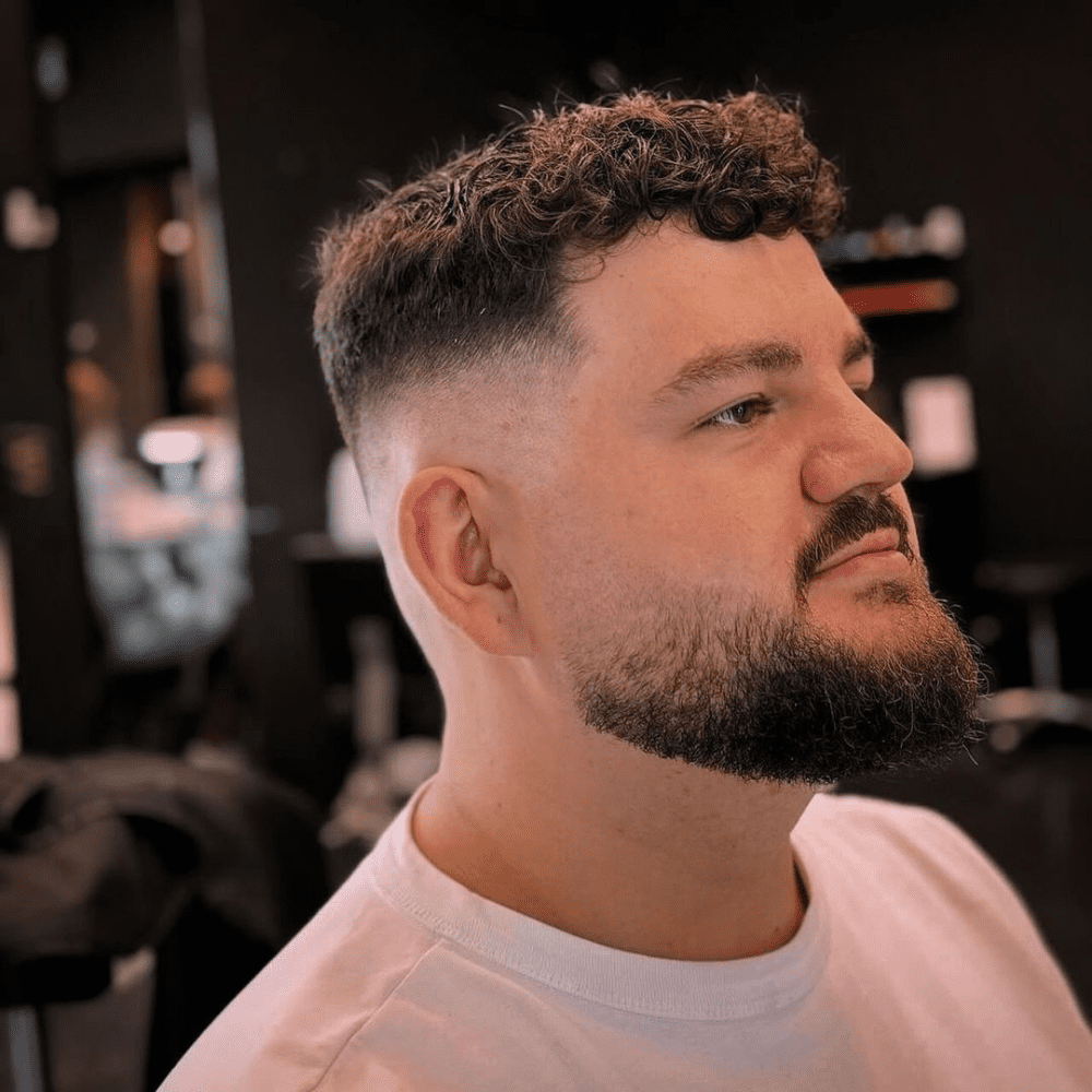 Side profile of a man with a curly high fade haircut, featuring tight curls on top that gradually fade into closely shaved sides. The haircut is complemented by a full, well-groomed beard, enhancing the overall stylish and modern look. The setting appears to be a barber shop, adding to the professional grooming atmosphere.