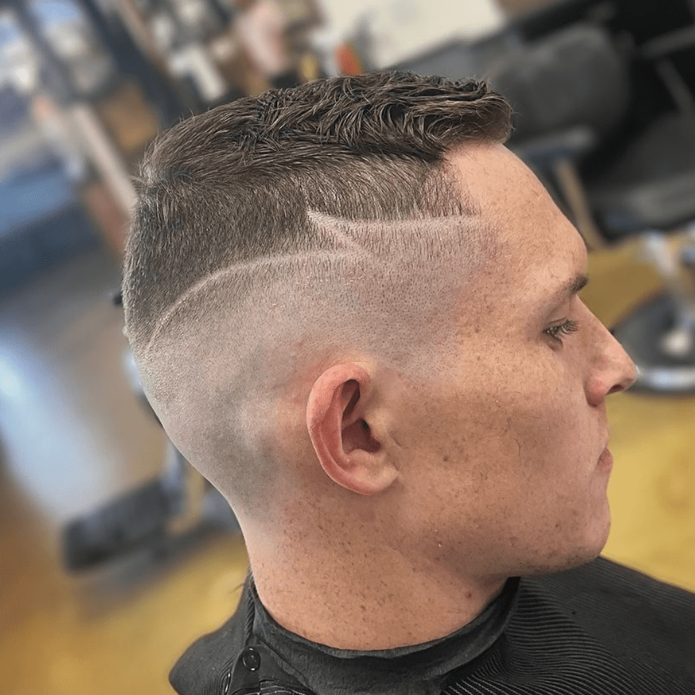 Side profile of a man with a high fade haircut featuring a unique design shaved into the closely shaved sides. The hair on top is slightly longer and textured, while the high fade incorporates a stylish, angular line design that adds a creative and personalized touch. The setting appears to be a barber shop, highlighting the professional and artistic grooming of the hairstyle.