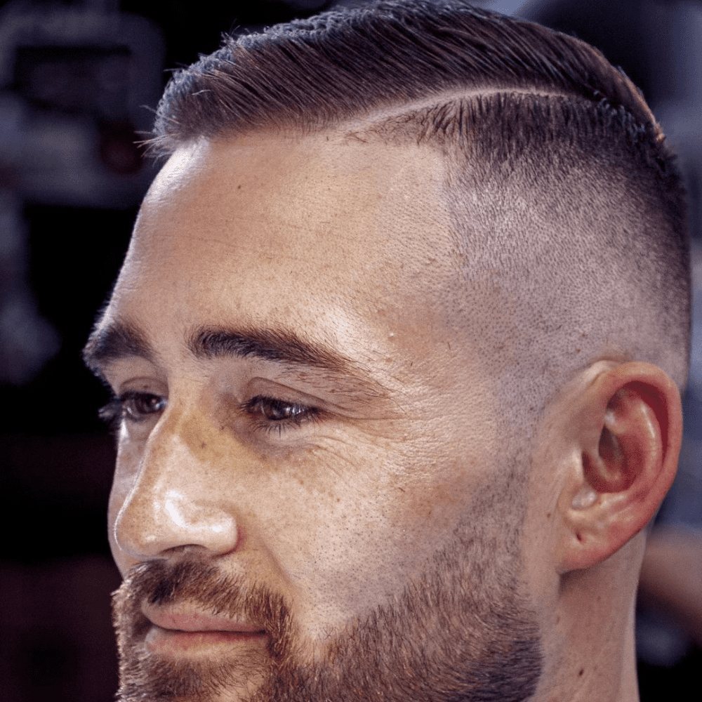 Close-up of a man with a high fade haircut featuring a defined part. The longer hair on top is neatly combed to one side, separated by a precise part line, while the sides are closely shaved with a high fade. The look is complemented by a short, well-groomed beard, giving a polished and professional appearance.