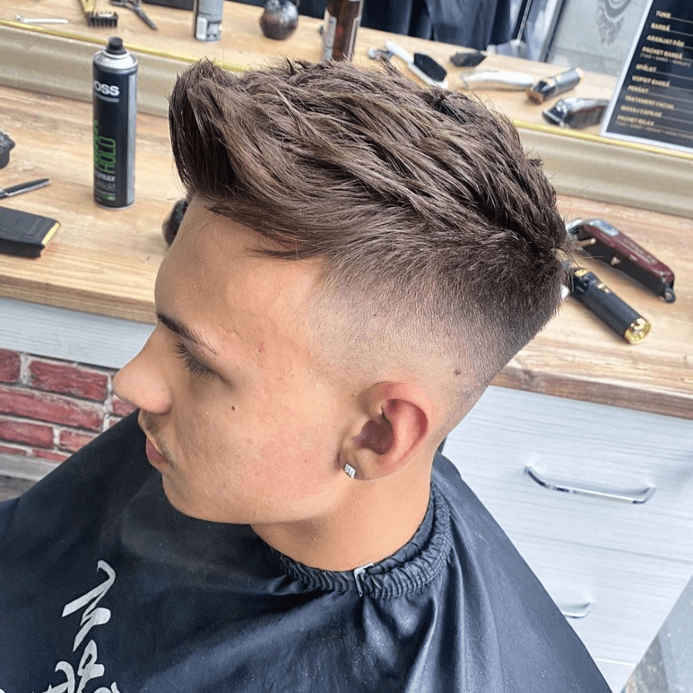 Side profile of a young man with a high fade haircut featuring a quiff, where the longer hair on top is styled upwards and back, creating volume and texture. The sides are neatly faded high, providing a sharp contrast that emphasizes the stylish quiff. The setting appears to be a barber shop, with various grooming tools visible in the background.