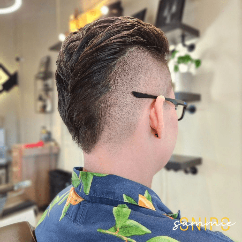 Rear view of a man with an Inverted Mohawk Fade haircut. The hairstyle features a unique twist on the traditional Mohawk, with the hair in the center shaved short and longer hair on the sides. The sides are styled back, creating a smooth, sleek look, while the high fade adds a clean, sharp contrast. The man is wearing glasses and a blue shirt with a colorful pattern, seated in a modern barbershop with stylish decor. The Inverted Mohawk Fade offers a bold and unconventional style that stands out.