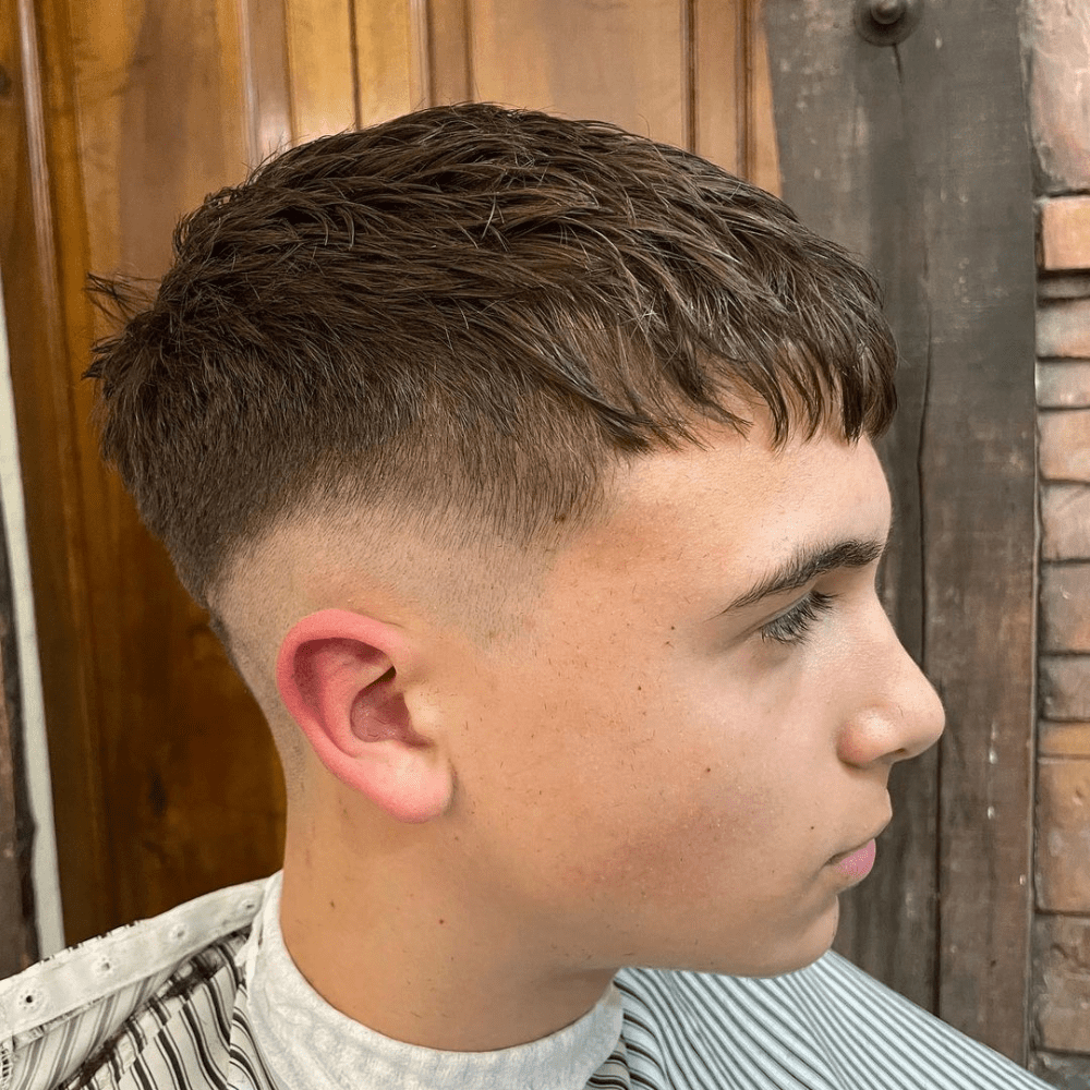 ide profile of a young man with an Ivy League haircut, featuring short, textured hair on top with a subtle fringe and a clean mid fade on the sides and back. The hairstyle provides a classic and sophisticated look, perfect for men seeking trendy yet refined haircuts