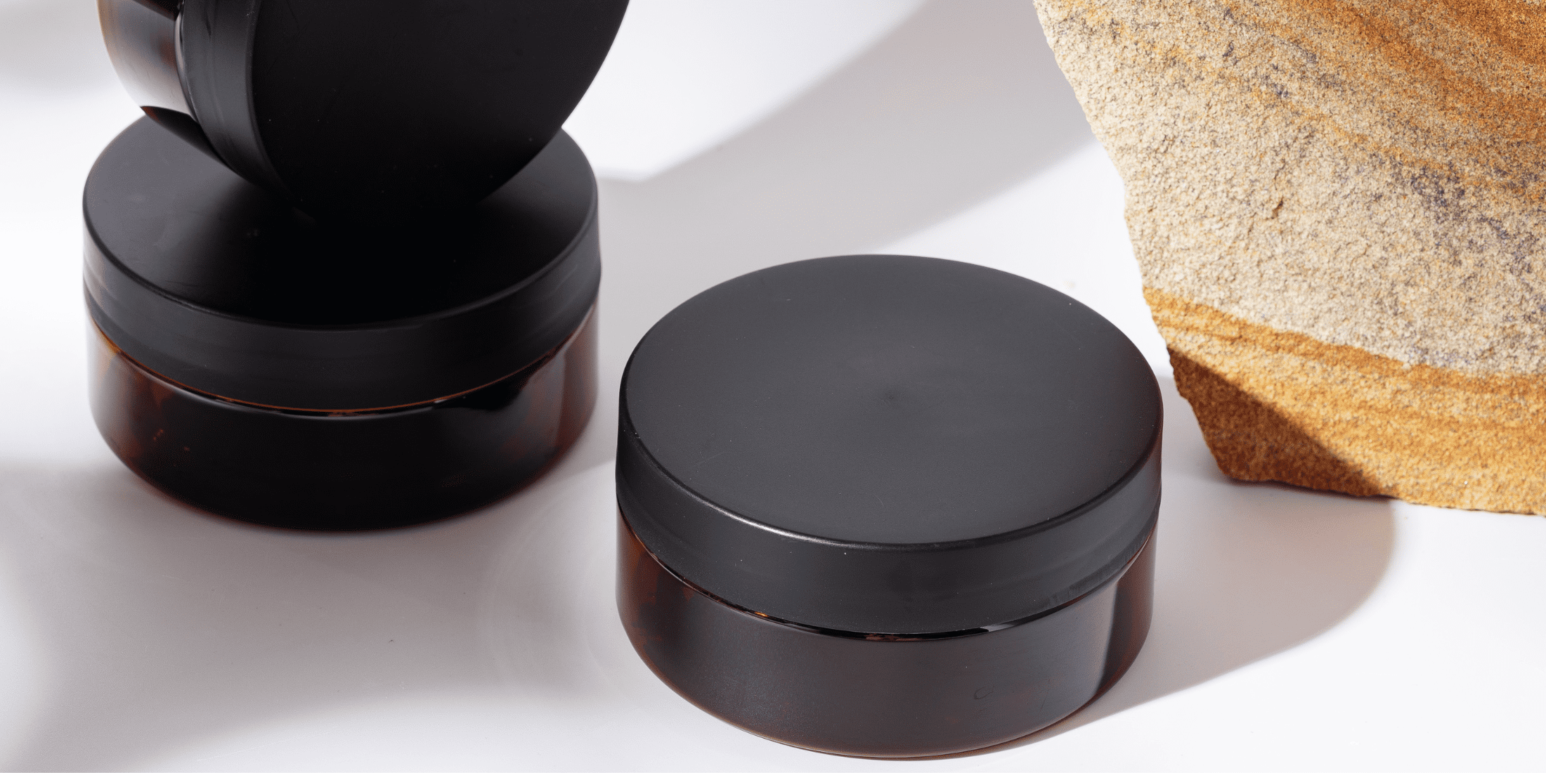 Jars of beard balm with sleek, black lids and amber-colored containers, placed on a white surface next to a textured stone. The image highlights the elegant and minimalist design of the beard balm packaging, perfect for the beard butter vs beard balm debate. Ideal for men looking for a product that provides light hold and control, showcasing the difference between beard balm and beard butter. This photo captures the essence of high-quality beard grooming products.