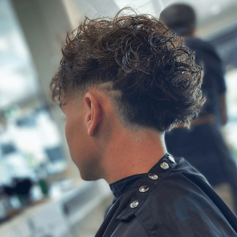 Side profile of a man with a Long Hair Burst Fade haircut, featuring voluminous and curly hair on top that gradually fades down the sides and back. The long curls add texture and movement, while the burst fade creates a clean and stylish transition. The fade starts high, near the temples, and tapers down, highlighting the contrast between the long top and the closely cropped sides. The setting is a modern barbershop, with a blurred background that keeps the focus on the expertly styled haircut. This long hair burst fade is perfect for those who want a trendy and eye-catching look.