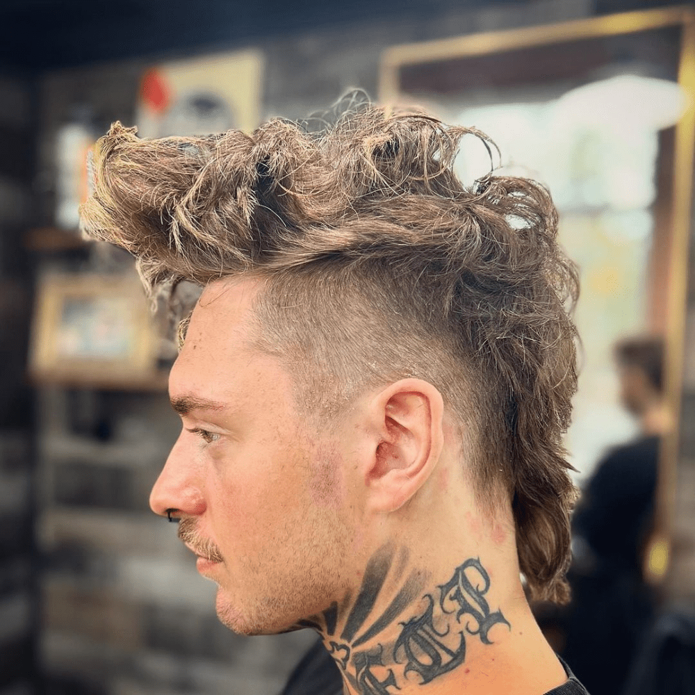 Side profile of a man with a Long Mohawk Fade haircut. The hairstyle features long, textured hair on top styled into a voluminous Mohawk, with the sides shaved into a high fade for a sharp contrast. The Mohawk extends down the back, adding length and dramatic flair. The man has visible neck tattoos and a mustache, enhancing his edgy and unique look. The background shows an indoor barbershop setting, highlighting the modern and bold style of the Long Mohawk Fade haircut.