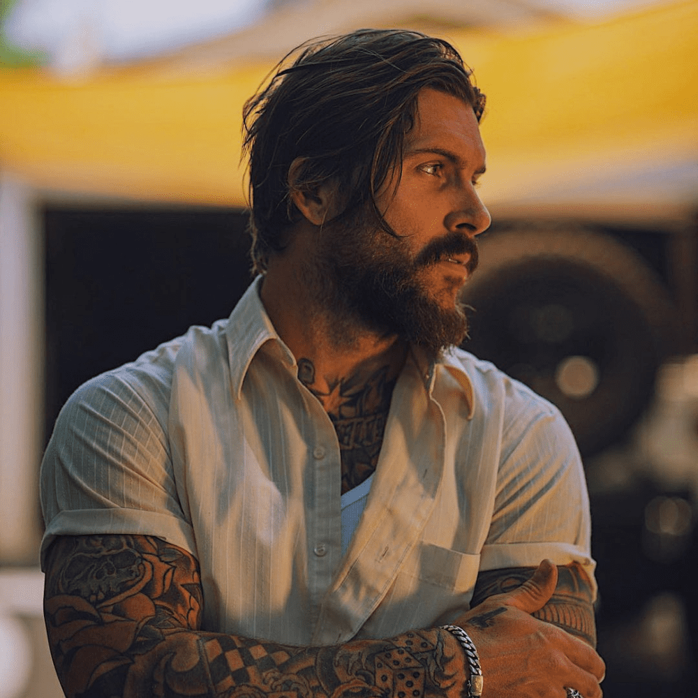 Man with a long slicked back haircut, showcasing a rugged yet polished look, paired with a full beard and visible tattoos. He is wearing a casual white shirt and standing in an outdoor setting. This image captures the essence of a modern, laid-back variation of the slicked back hairstyle, perfect for men with long hair in 2024 seeking a stylish and effortless appearance.