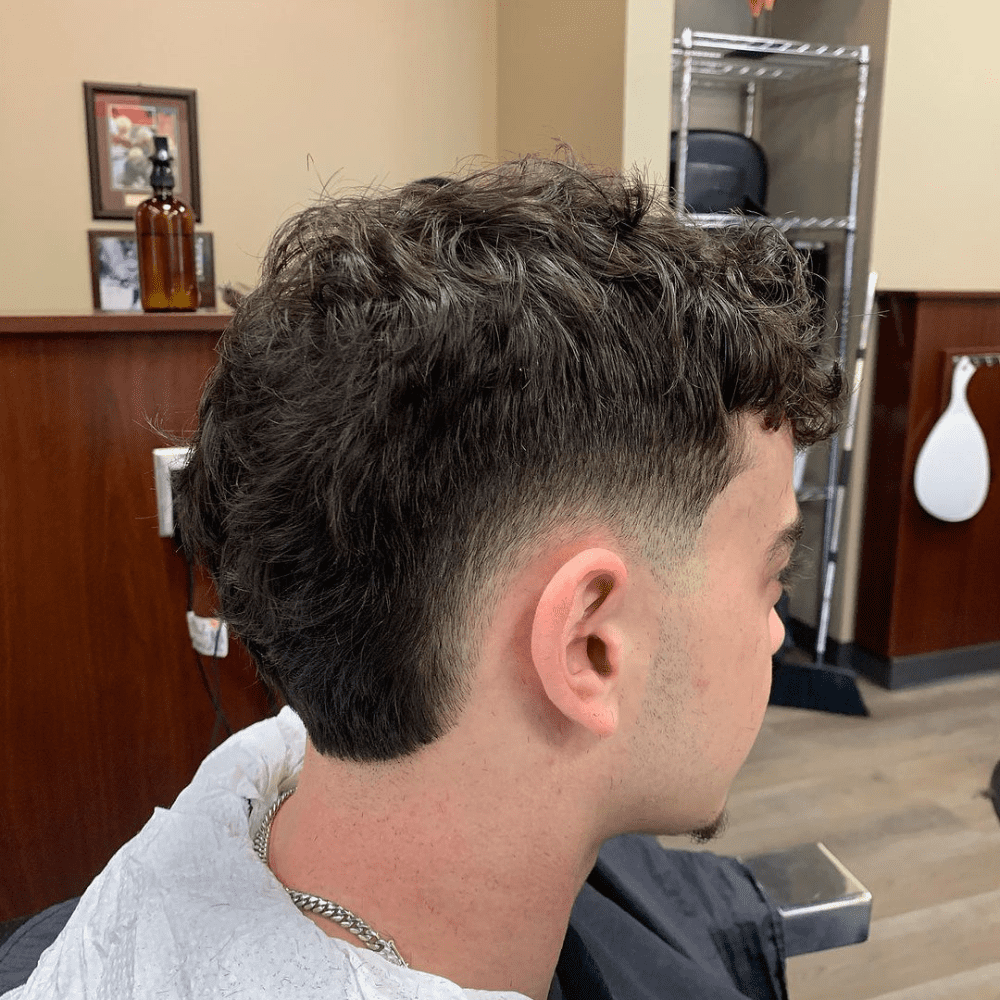 Side profile of a man with a Low Burst Fade haircut, showcasing a subtle and sophisticated fade that starts just above the ears. The hairstyle features thick, wavy hair on top, gradually tapering down to shorter lengths at the sides and back. This low burst fade provides a clean and polished look, suitable for both professional and casual settings. The background includes a barber shop setting with wooden panels and grooming tools, emphasizing the fresh, well-groomed style.