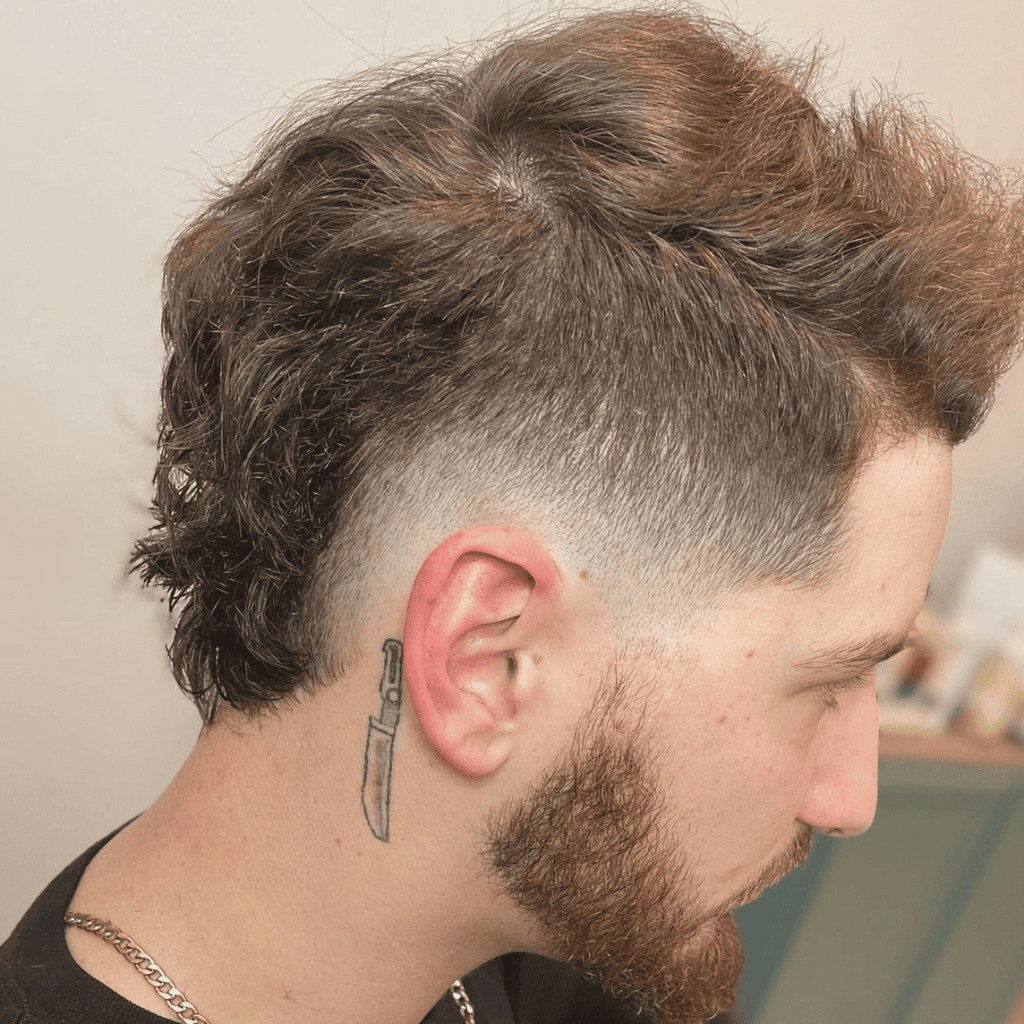 Side profile of a man with a low fade haircut, textured hair on top, and a gradual fade around the ear. The style is complemented by a beard and a knife tattoo behind the ear, creating a distinctive and edgy appearance.