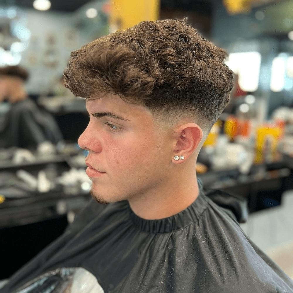 Side profile of a young man with a neat low fade haircut, featuring curly brown hair on top. The fade starts just above the ear, creating a smooth and subtle transition. This image showcases the low fade style in the drop fade vs low fade comparison, highlighting its clean and classic appearance.