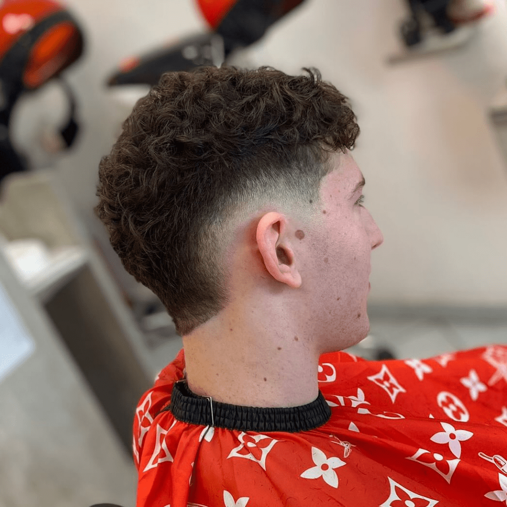 Side profile of a young man with a Low Fade Mohawk haircut. The hairstyle features curly hair on top, styled into a Mohawk with a gradual low fade on the sides. The fade starts lower on the head, creating a subtle transition between the longer curls on top and the shorter sides. The man is wearing a red barber cape with a white pattern, sitting in a barbershop setting. The Low Fade Mohawk highlights the natural texture of the curls while maintaining a clean and modern look.