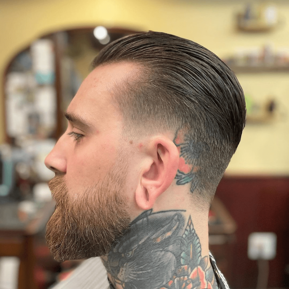 Side profile of a man with a low fade slicked back haircut, featuring neatly trimmed sides that gradually blend into the longer, slicked back top. The man has a well-groomed beard and visible neck tattoos, showcasing a modern and edgy variation of the slicked back hairstyle. This image highlights a stylish and polished look ideal for men seeking a contemporary yet classic haircut in 2024.