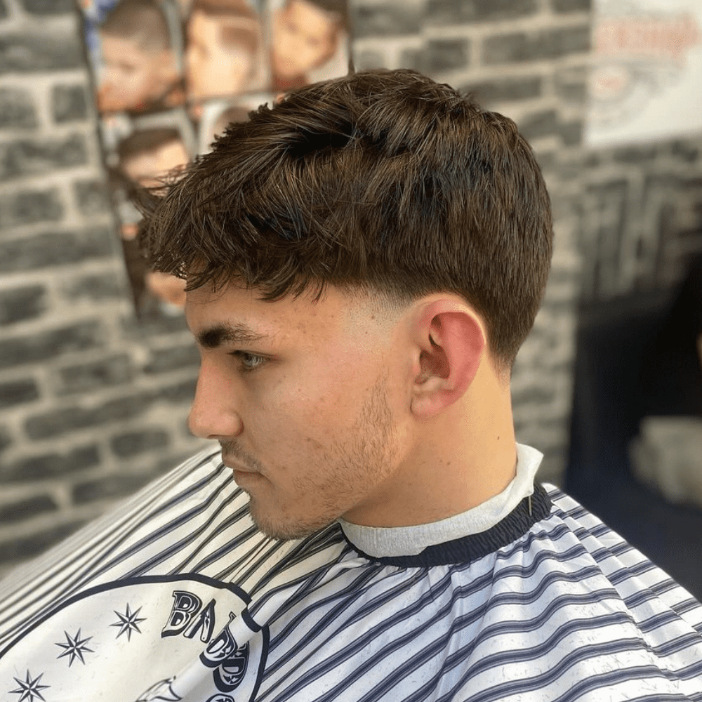 Side profile of a young man with a Low Taper Fade haircut, featuring short, textured hair on top with a subtle taper fade starting just above the ears. The hairstyle offers a refined and understated look, making it an ideal choice for men seeking trendy yet classic fade haircuts