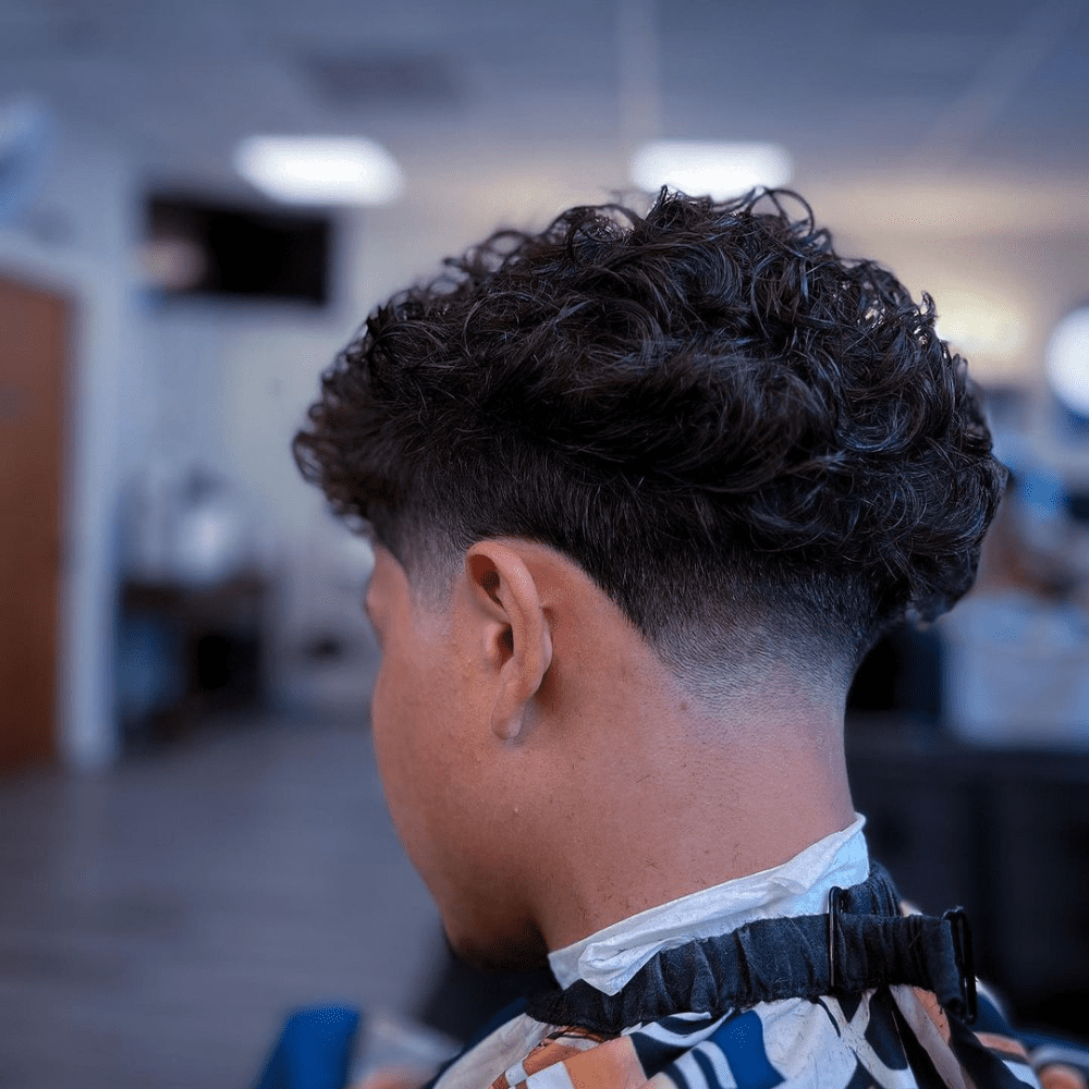 A close-up of a young man sporting a stylish mid taper haircut. The haircut features a gradual fade starting around the midpoint between the ears and temples, blending seamlessly into his naturally curly hair on top. This image highlights the key differences in the low taper vs mid taper debate, showcasing the mid taper's more noticeable transition and trendy appearance.