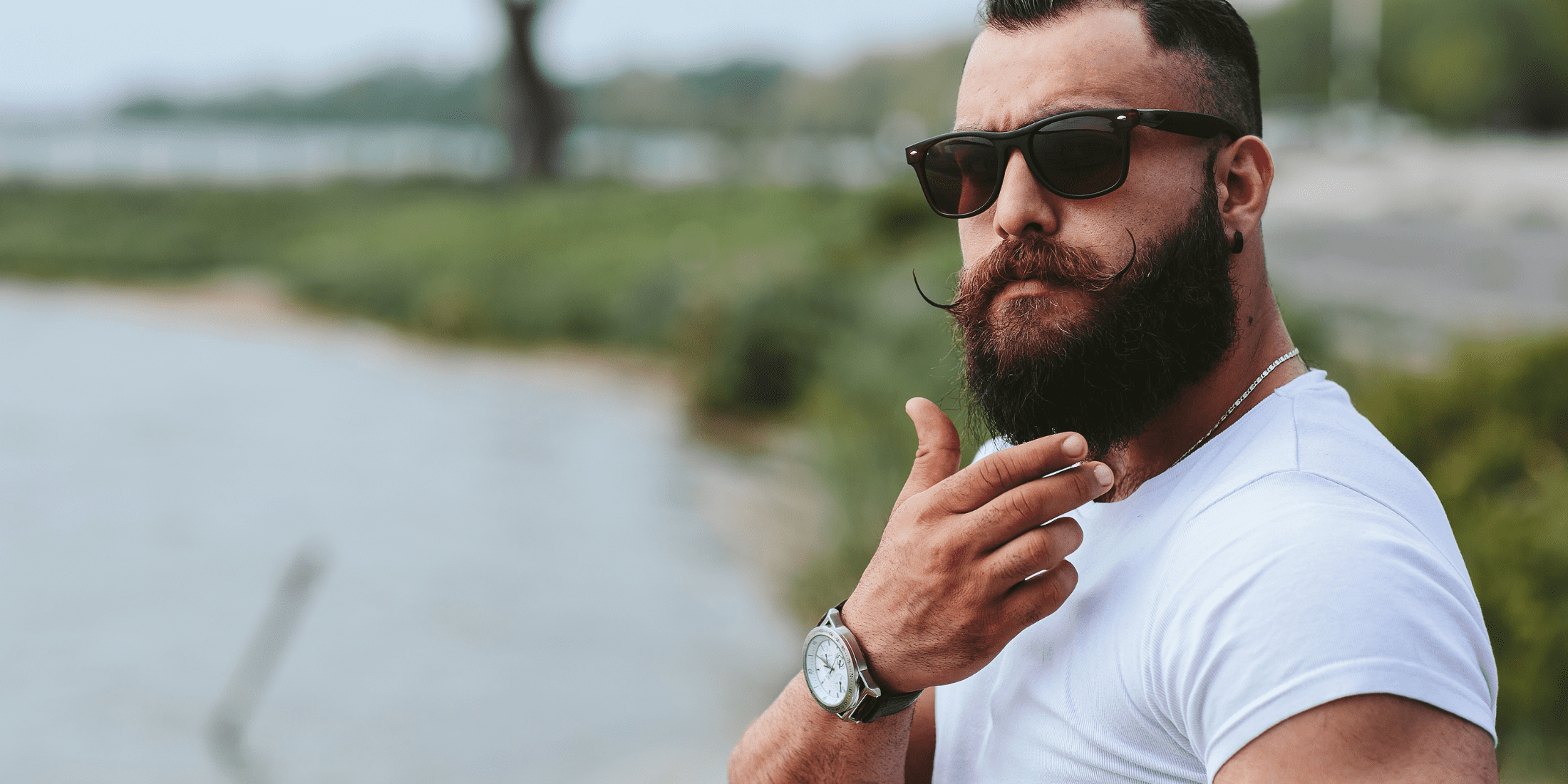 Man with a well-groomed beard and sunglasses standing by a scenic lakeside, exemplifying a healthy, dandruff-free beard.