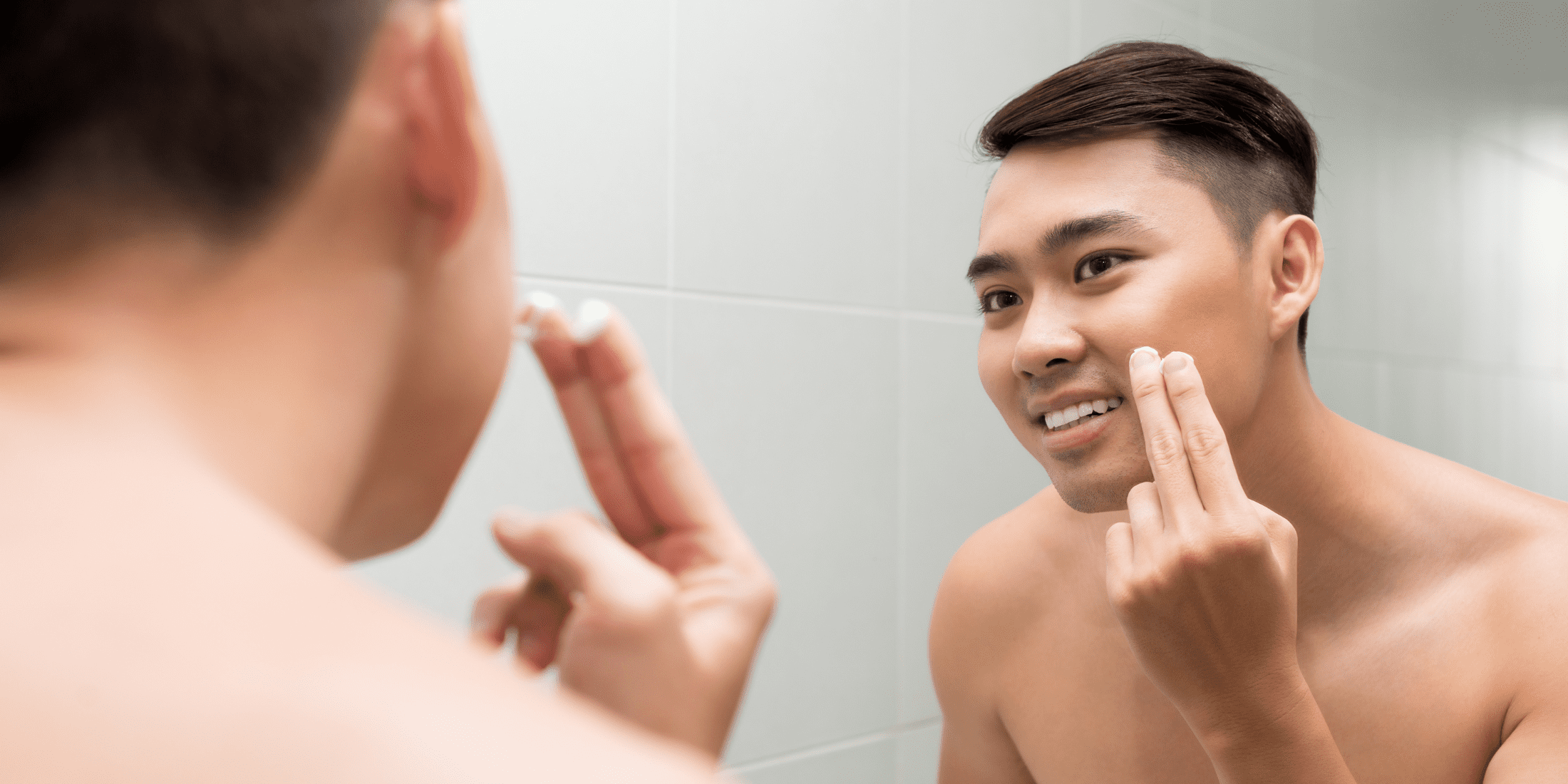 A young man with short, dark hair is standing in front of a bathroom mirror, smiling as he applies aftershave to his face. The setting is modern with a tiled wall, and the man appears to be enjoying his grooming routine. This image captures the moment of self-care, illustrating the use of aftershave in a daily skincare regimen, emphasizing the difference in aftershave vs. cologne usage.