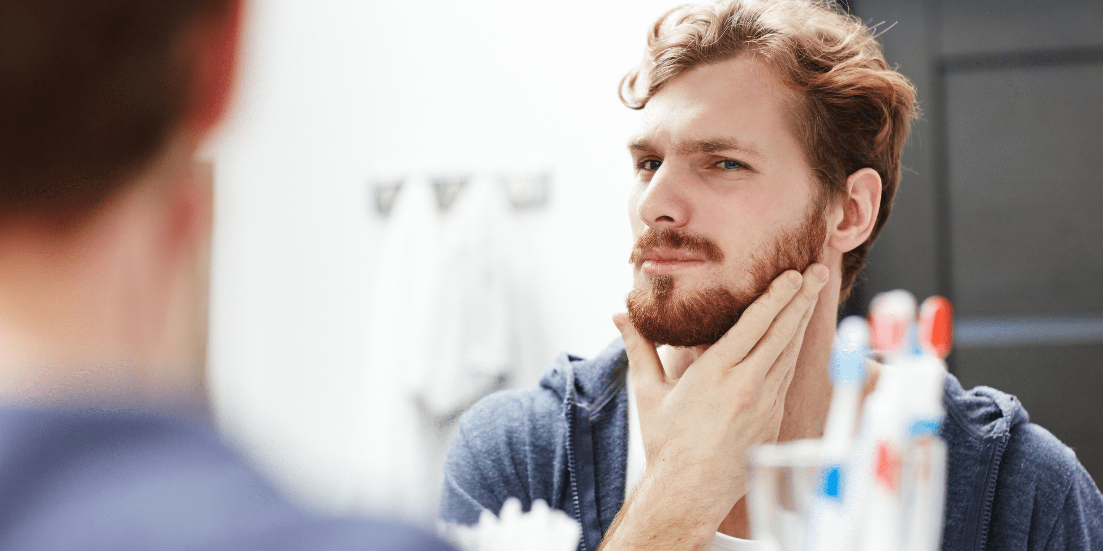 A man standing in front of a bathroom mirror, applying beard balm to his beard. The image captures him using his hands to evenly distribute the balm, demonstrating how to use beard balm for optimal beard care.