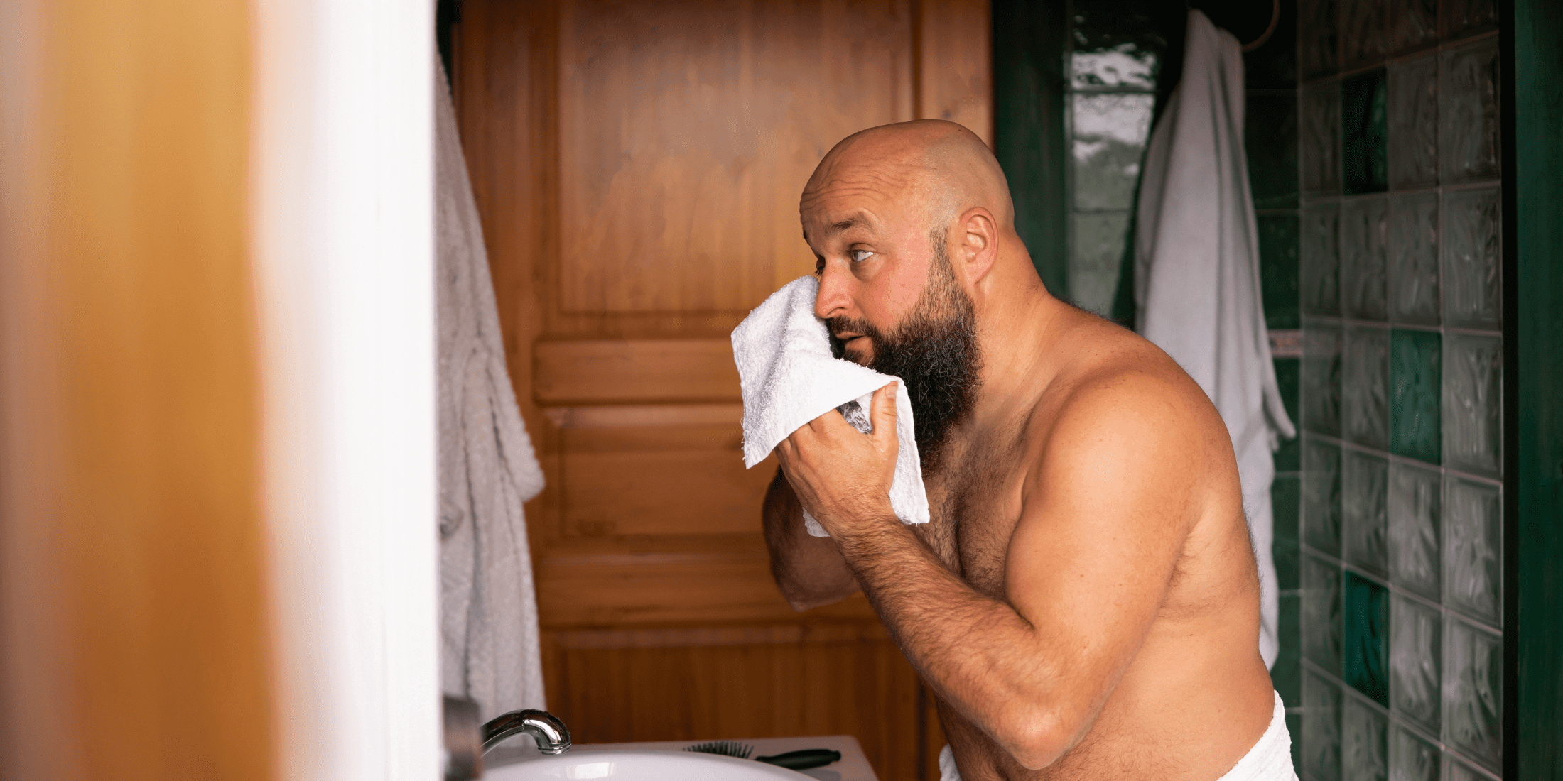 A man standing in front of a bathroom mirror, towel drying his beard after a shower, preparing to use beard balm. The image captures the initial steps of how to use beard balm, emphasizing the importance of starting with a clean and damp beard.