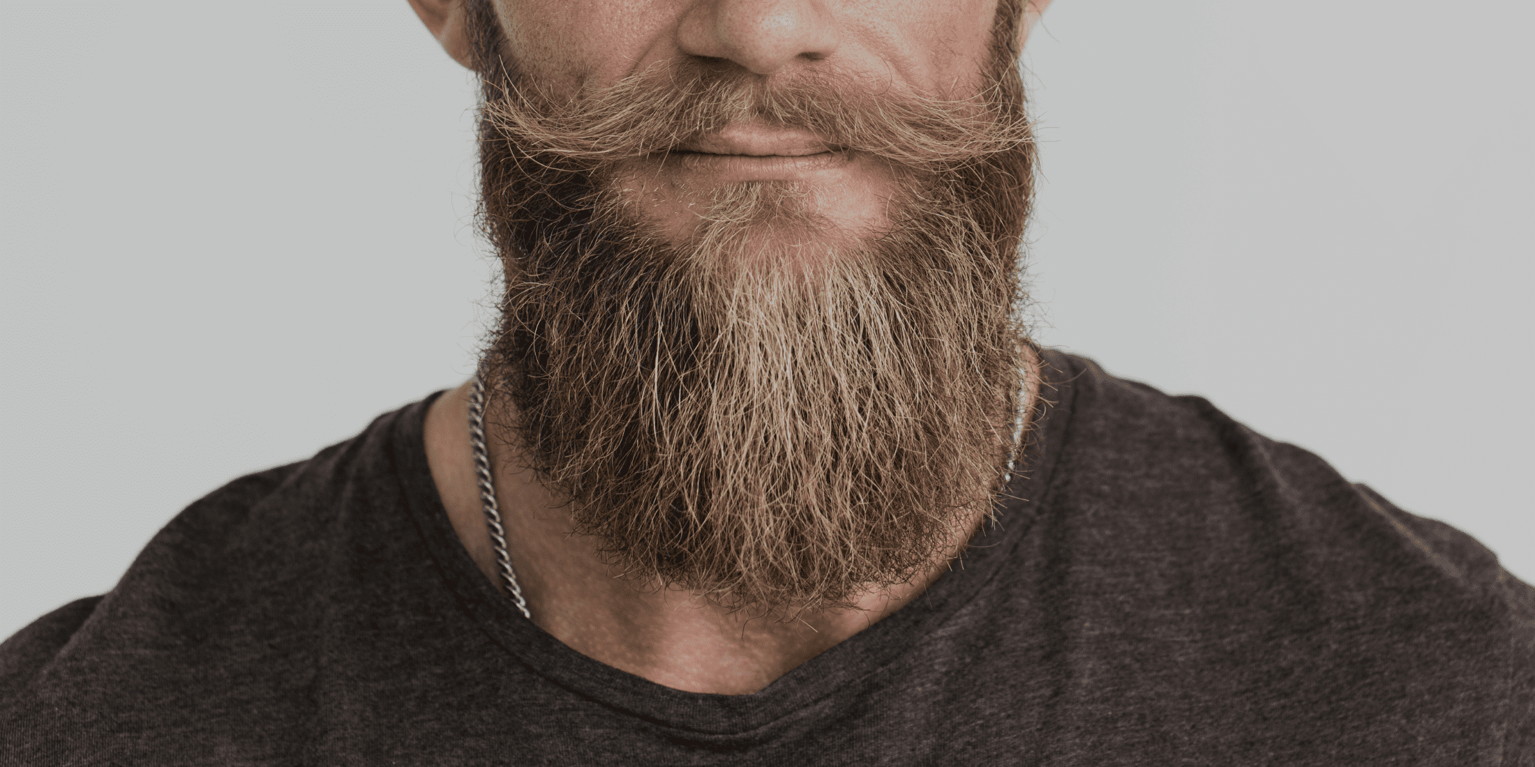 A close-up image of a man with a well-groomed, full beard and styled mustache. The beard appears thick and textured, showcasing a mix of coarse and softer hair. The man is wearing a dark t-shirt and a silver chain necklace, and his facial features, including his lips and part of his nose, are visible. This image highlights beard texture and grooming, making it ideal for discussing beard care routines and tips on how to make your beard soft.