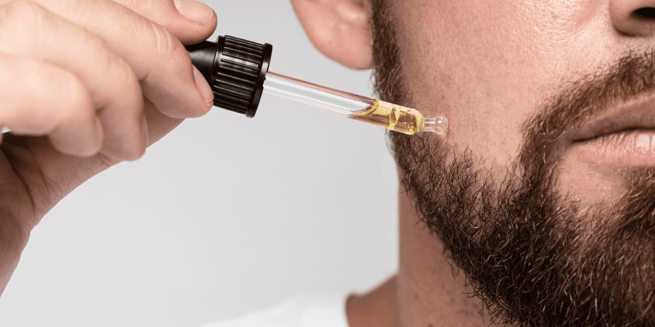 A close-up image of a man applying beard oil to his beard using a dropper. The focus is on the dropper releasing golden oil onto the side of his face near his well-groomed beard. The man is wearing a white shirt, and the background is neutral, highlighting the beard care process. This image emphasizes the importance of using beard oil to maintain a soft, healthy beard.