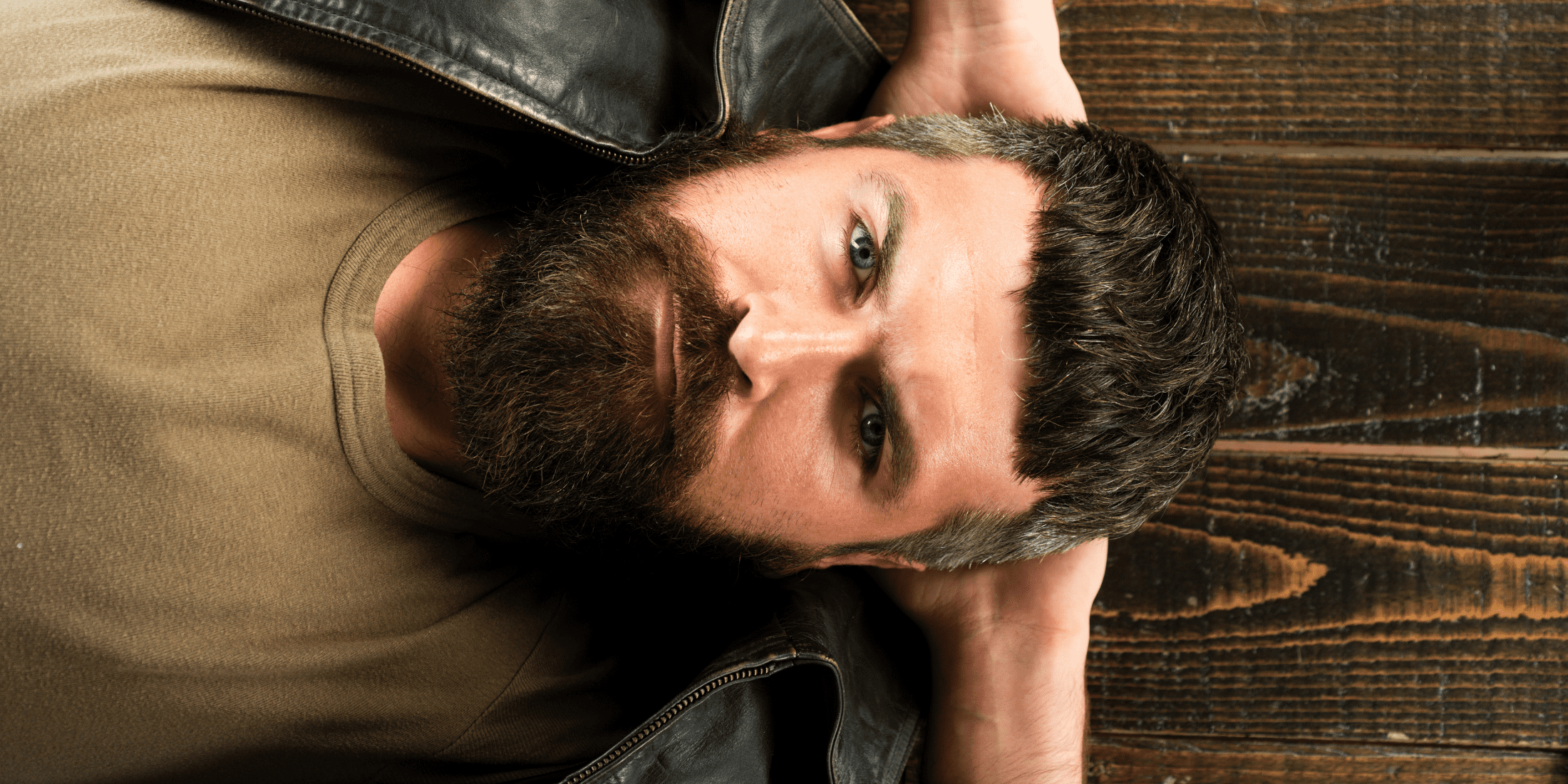 A man with a thick, well-groomed beard lies on his back on a wooden surface, looking contemplatively upward. He is wearing a brown shirt and a black leather jacket, with his hands resting behind his head. The image captures a thoughtful expression, possibly pondering the question, Why did my beard stop growing?