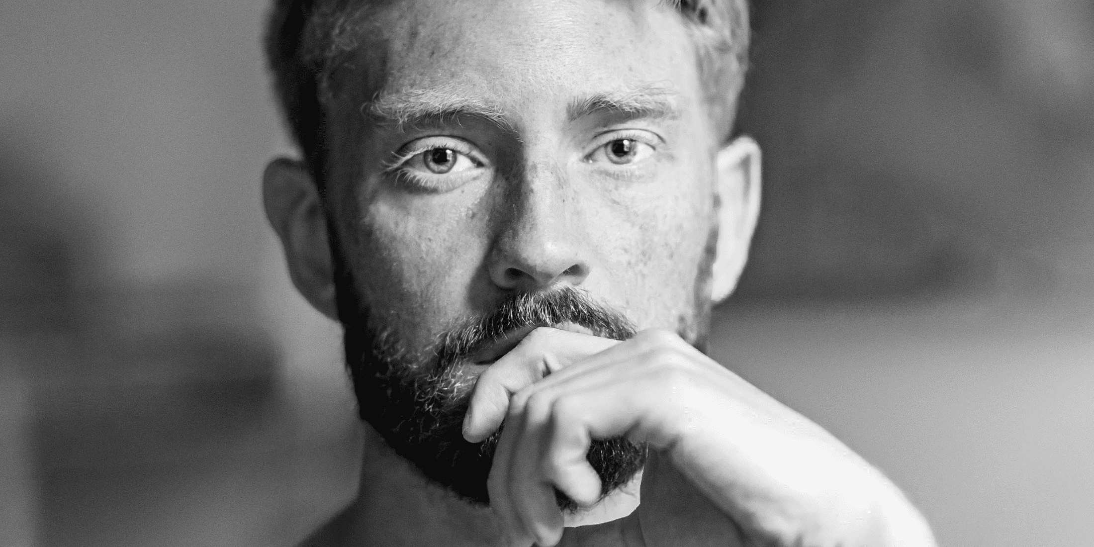 A black-and-white close-up image of a man with a well-groomed beard. He rests his chin on his hand and looks thoughtfully into the camera. The contemplative expression suggests he might be pondering a question like, Why did my beard stop growing? The background is softly blurred, keeping the focus on the man's face and introspective mood.