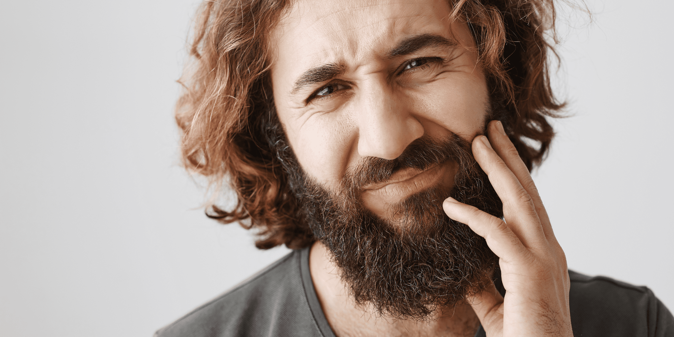 Portrait of a man with dry facial hair, showing discomfort and touching his beard, highlighting the rough texture and irritation associated with dry beard conditions.