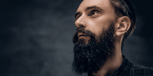 A close-up, side-profile portrait of a man with a full, frizzy beard, gazing upwards thoughtfully. The lighting emphasizes the texture of his thick beard and the sharp features of his face. 