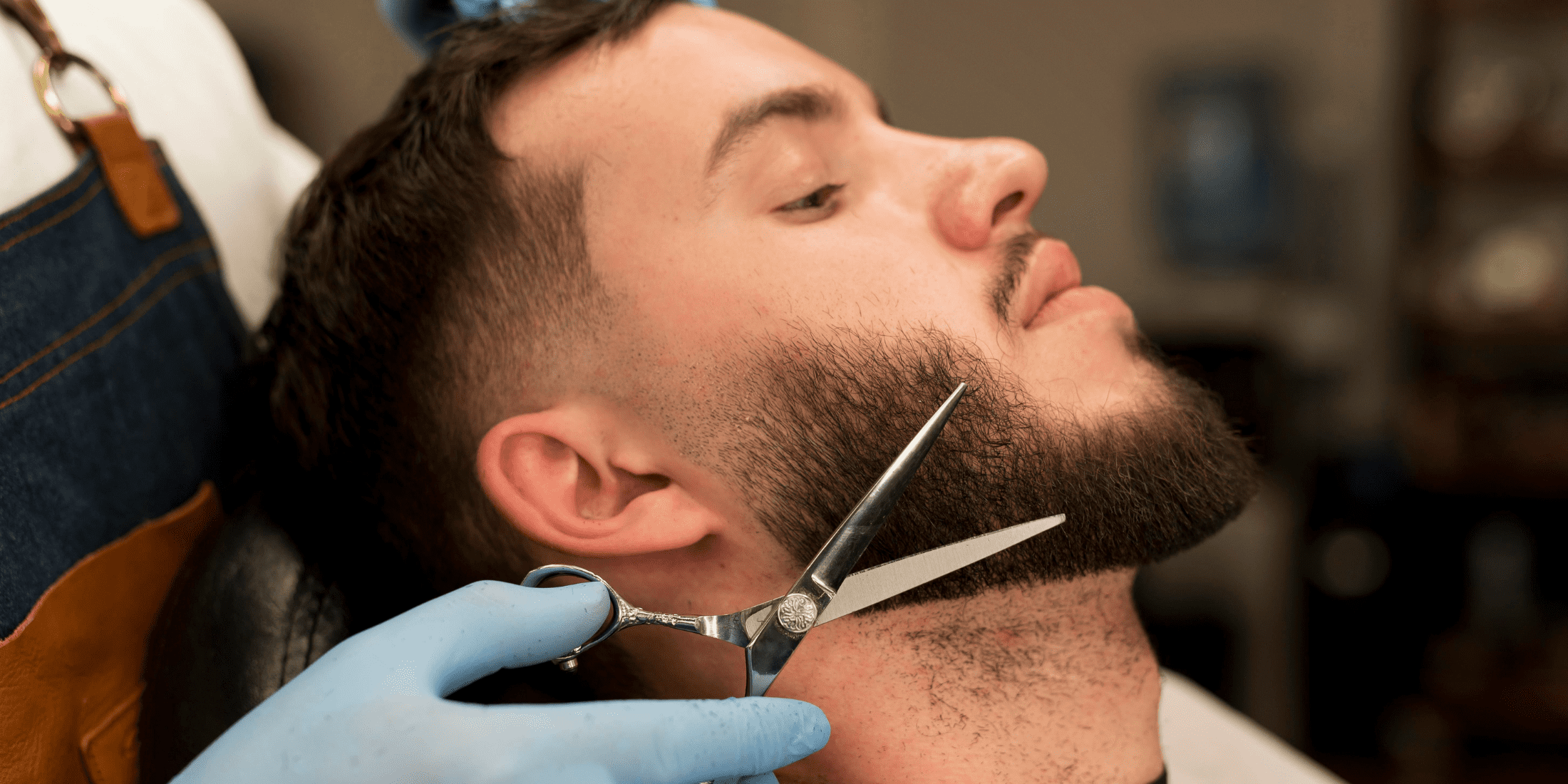 A close-up image of a man receiving a beard trim at a barbershop. The man is reclining comfortably while a barber, wearing blue gloves, carefully trims his beard with professional scissors. The focus is on the precision of the trim, highlighting the barber's skill in shaping and maintaining the beard. This image emphasizes the importance of regular trimming to prevent and manage beard split ends and keep the beard looking well-groomed and healthy.