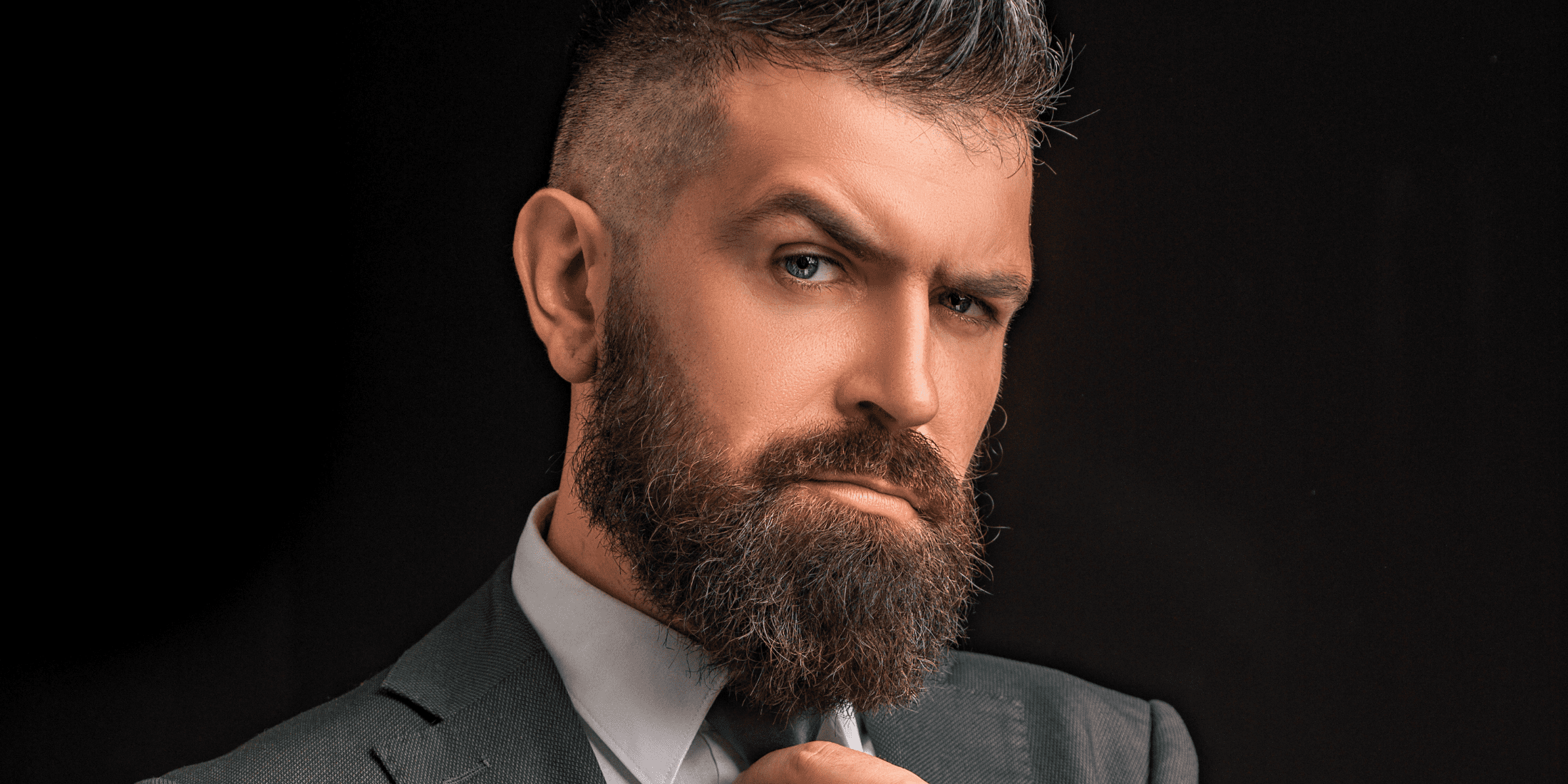 A close-up image of a well-groomed man with a full, thick beard and styled short hair. He has an intense, confident expression, with one hand adjusting his tie. The background is dark, making his beard and facial features stand out prominently. This image highlights the potential presence of beard split ends, emphasizing the importance of proper beard care and grooming