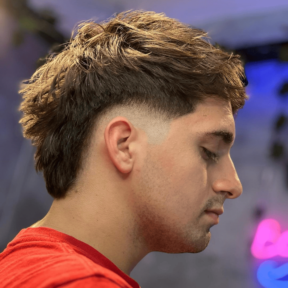 Side profile of a young man with a Medium Mohawk Fade haircut. The hairstyle features medium-length hair on top, styled into a Mohawk with a high fade on the sides. The fade creates a sharp contrast with the longer hair, which has a textured, slightly tousled look. The man is wearing a red shirt, and the background suggests an indoor setting with soft, colorful lighting. The Medium Mohawk Fade showcases a balanced and stylish appearance, combining modern edginess with a versatile length.