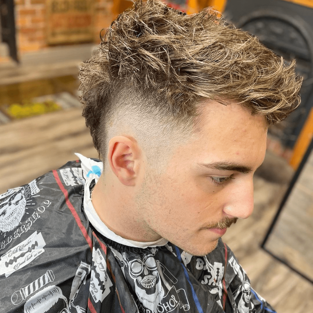 Side profile of a young man with a Messy Mohawk Fade haircut. The hairstyle features a textured Mohawk with a deliberately tousled and messy appearance on top, combined with a high fade on the sides. The fade creates a clean contrast with the disheveled top, enhancing the edgy and casual look. The man is wearing a black barber cape with various designs and is seated in a barbershop with a wooden floor and brick walls, showcasing the trendy and relaxed style of the Messy Mohawk Fade.