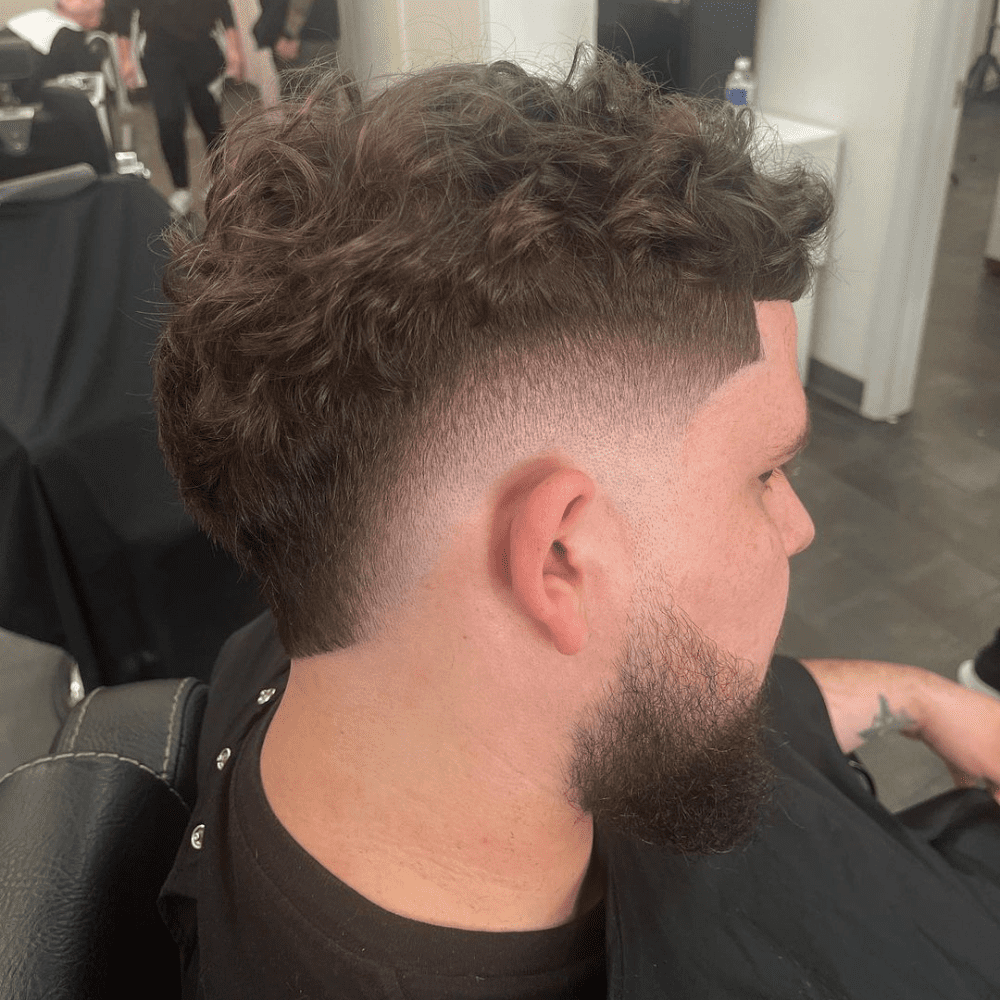 Side profile of a man with a Mid Burst Fade haircut, showcasing thick, curly hair on top that gradually fades to a shorter length at the sides and back. The fade starts around the temple area, providing a balanced and stylish look. The haircut is complemented by a neatly trimmed beard that seamlessly blends with the fade, creating a cohesive and well-groomed appearance. The setting is a modern barbershop, highlighting the fresh and expertly styled haircut. This mid burst fade is perfect for adding structure to curly hair while maintaining a trendy and fashionable look.