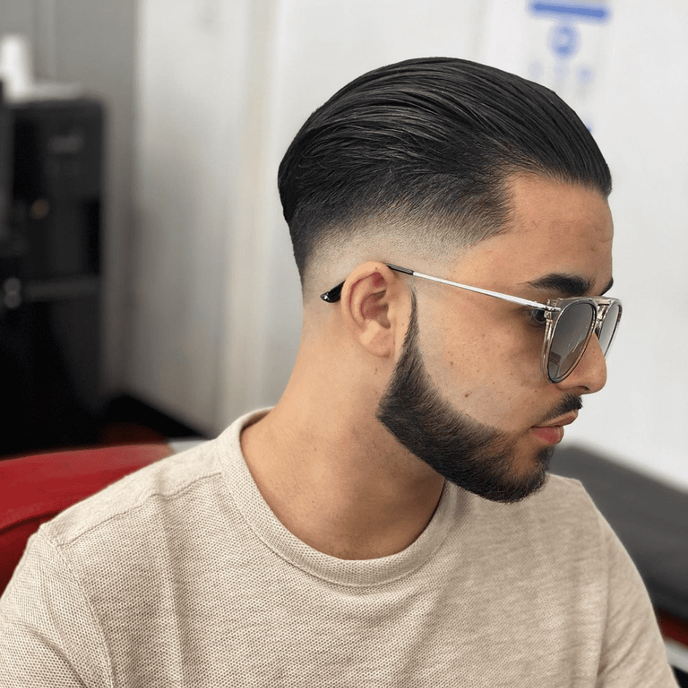 Side profile of a man with a mid fade slicked back haircut, featuring a smooth transition from the faded sides to the longer, slicked back top. He is wearing sunglasses and has a sharply defined beard, adding to the modern and stylish look. This image showcases a clean and contemporary variation of the slicked back hairstyle, perfect for men in 2024 seeking a trendy yet polished appearance.