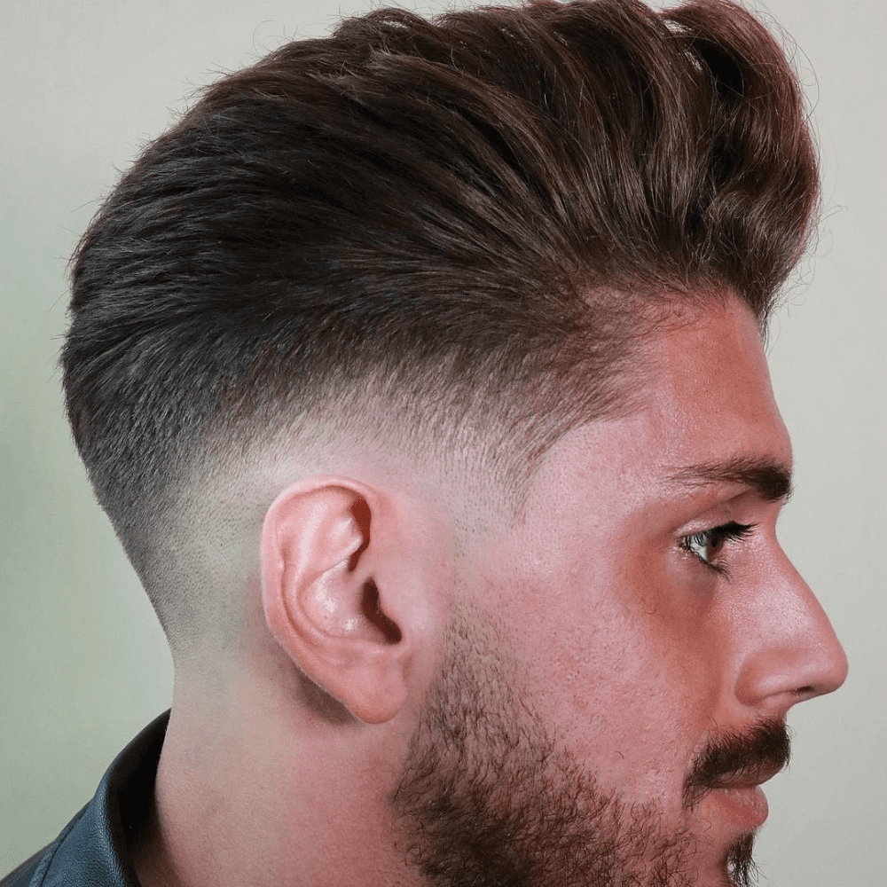 A side profile of a man showcases a modern pompadour haircut. The hair on top is voluminous, styled upwards and back, creating a sleek and full appearance. The sides are tapered into a smooth fade, blending seamlessly into the longer top. The man has a well-groomed beard that complements the sharp, clean lines of the haircut. The image highlights the contrast between the short, faded sides and the lush, styled top, exemplifying a contemporary take on the classic pompadour.