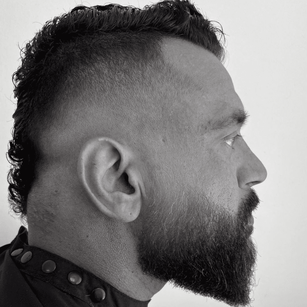 Side profile of a man with a Mohawk Fade haircut paired with a full beard. The hairstyle features a high fade on the sides, with a textured Mohawk extending from the front to the back. The full beard complements the edgy Mohawk, creating a rugged yet stylish look. The man is wearing a dark barber cape, and the background is a plain white wall, highlighting the sharp contrast and clean lines of the Mohawk Fade with Beard. This combination showcases a bold and masculine style.