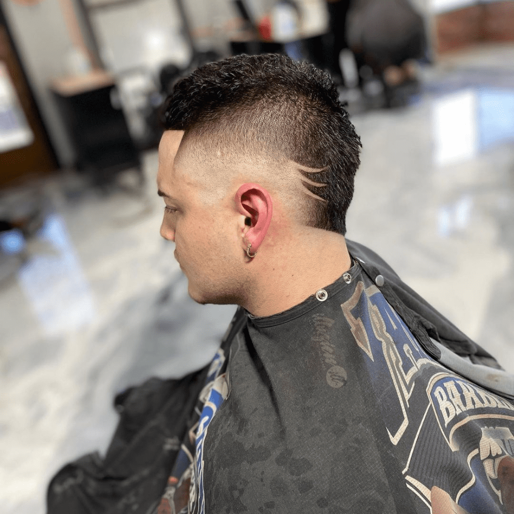 Side profile of a man with a Mohawk Fade haircut featuring a unique design. The hairstyle showcases a high fade on the sides, with the Mohawk extending from the front to the nape. The sides feature a creative shaved design, adding a personalized and artistic touch to the look. The man is seated in a barbershop, wearing a black barber cape. The Mohawk Fade with Design combines modern edginess with artistic elements, making it a bold and stylish choice.