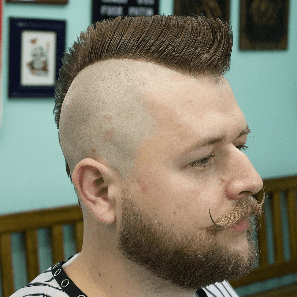 A man with a well-groomed beard and a neatly styled Mohawk, featuring shaved sides and a prominent strip of hair down the center, sits in a barber chair. The Mohawk showcases a sleek and polished look, highlighting the dramatic difference in the Mohawk vs Faux Hawk debate.