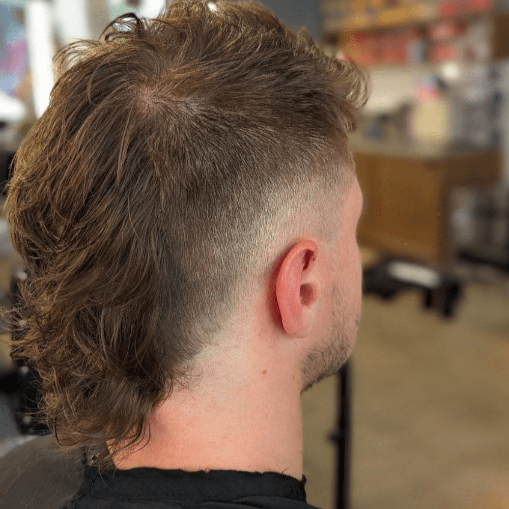 Rear view of a man with a Mohawk Mullet Fade haircut. The hairstyle features longer hair on the top and back, blending the edgy Mohawk with the retro mullet style. The sides are faded, creating a sharp contrast with the longer hair on top and at the back. The man is seated in a barbershop, showcasing the unique combination of a Mohawk and mullet with a clean, modern fade.