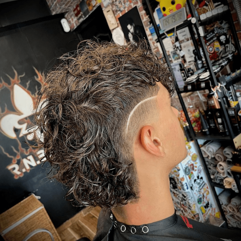 Side profile of a man with a Mullet Burst Fade haircut, featuring long, curly hair on top and back that transitions into a burst fade on the sides. The fade starts high at the temples and tapers down smoothly, with a distinctive shaved line adding a stylish and edgy detail. The longer curls flow towards the back, creating the classic mullet shape with a modern twist. The setting is a vibrant barbershop, filled with various grooming products and posters, highlighting the trendy and professional atmosphere. This mullet burst fade is perfect for men looking to combine retro and contemporary styles for a unique and fashionable look.