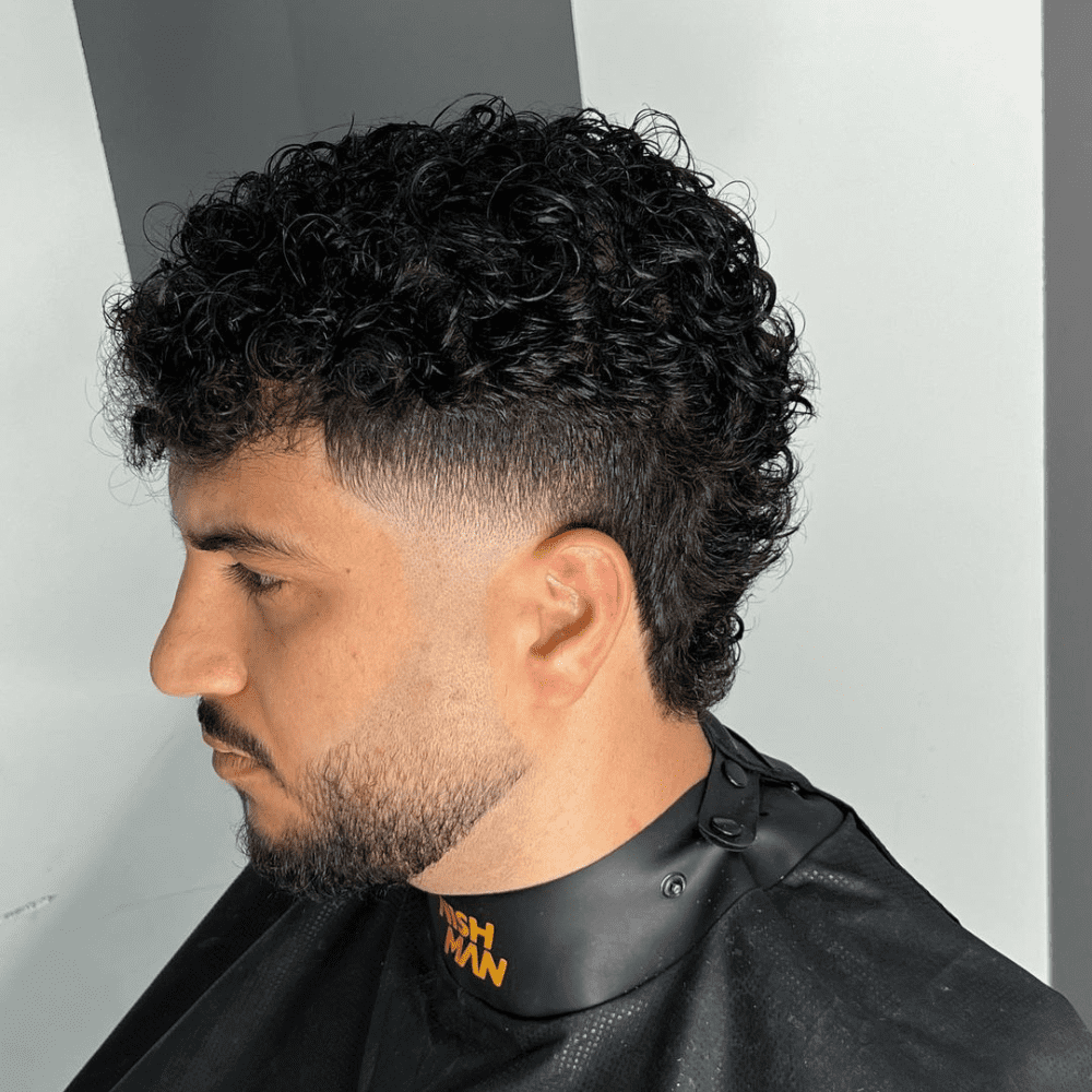 Man with a short fade haircut featuring a curly mullet fade. The hairstyle includes a clean fade on the sides and back, blending into the longer, curly hair at the top and back. This bold and trendy look is ideal for modern men's hairstyles in 2024, offering a unique and stylish appearance.