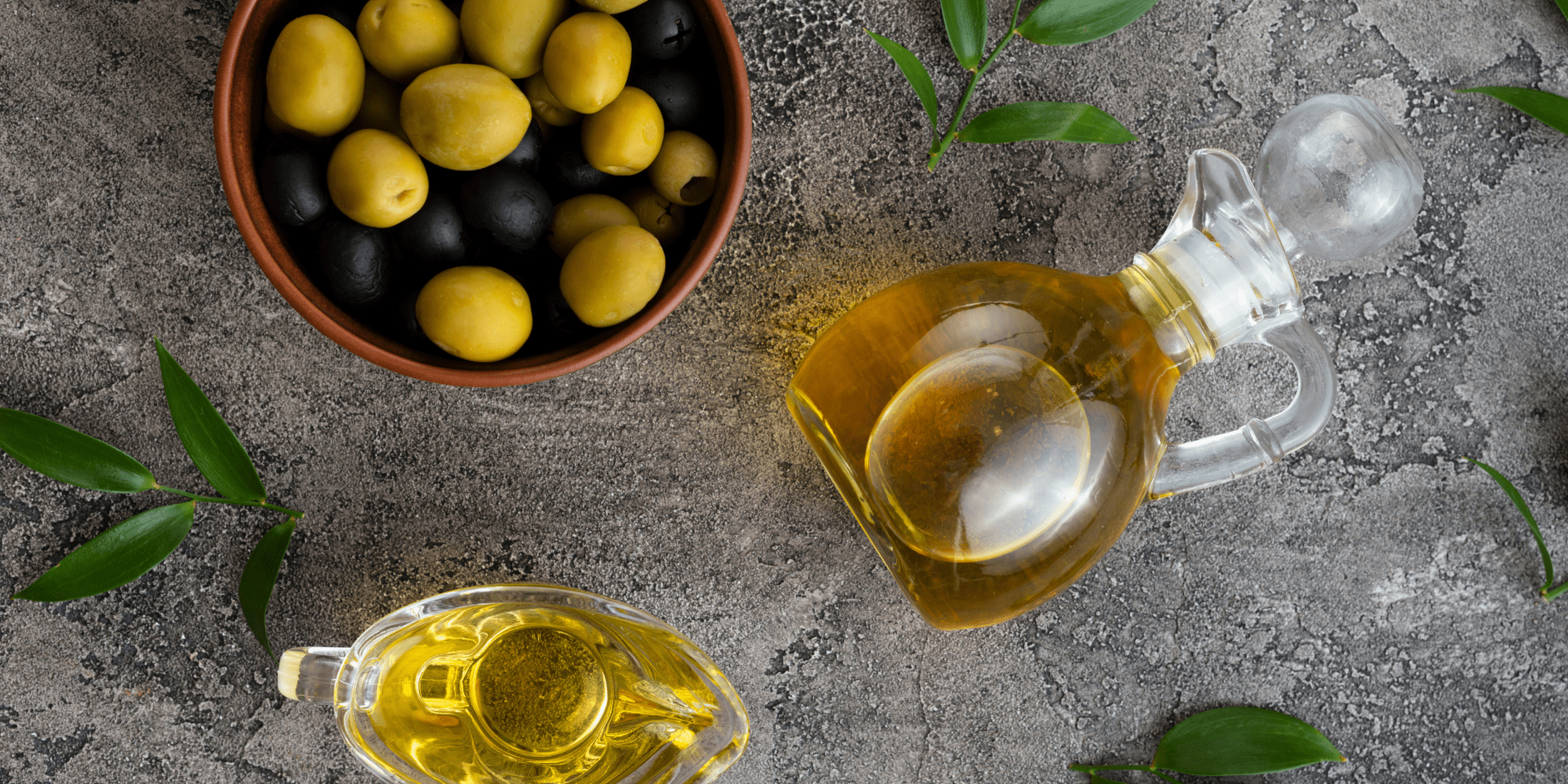 A top view of a bowl filled with green and black olives alongside two glass containers of olive oil, placed on a textured gray surface with green leaves scattered around, illustrating the natural ingredients used in olive oil for beard care.