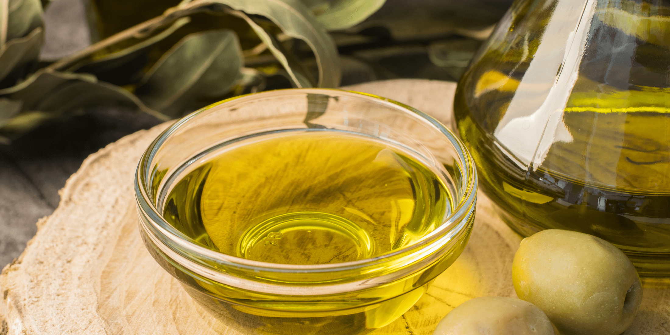 A close-up view of a small glass bowl filled with olive oil, placed on a wooden surface next to a larger glass container of olive oil and a couple of green olives, with olive branches in the background, showcasing the natural ingredients used in olive oil for beard care.