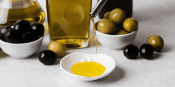 Olive oil being poured into a small dish, surrounded by bowls of green and black olives, with bottles of olive oil in the background, highlighting olive oil for beard care.