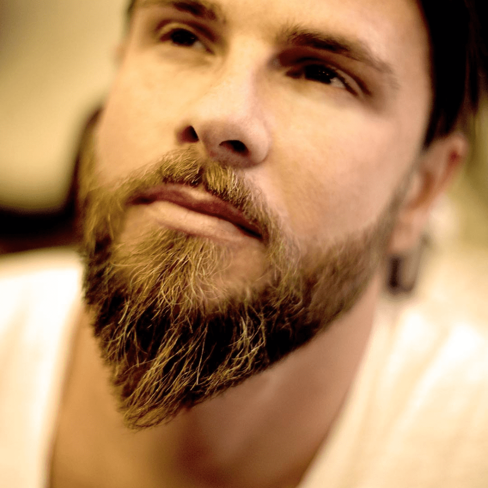 Close-up of a man with a pointed ducktail beard, showcasing the sharp taper and meticulous grooming of his facial hair, set against a blurred background that highlights the beard's defined shape and texture