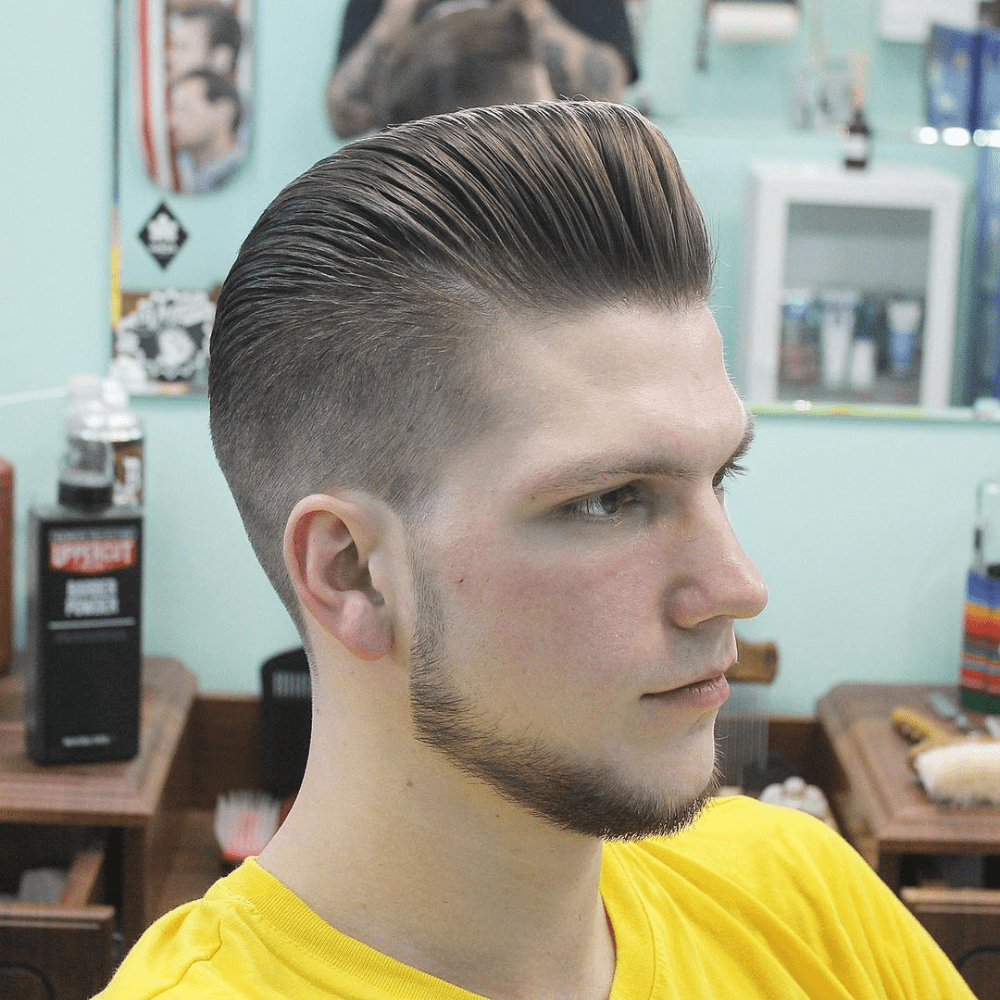 Side profile of a man with a Pompadour Fade haircut, featuring voluminous hair styled high and slicked back on top, paired with a clean fade on the sides and back. The hairstyle blends classic and modern elements, making it a stylish choice for men seeking trendy fade haircuts.