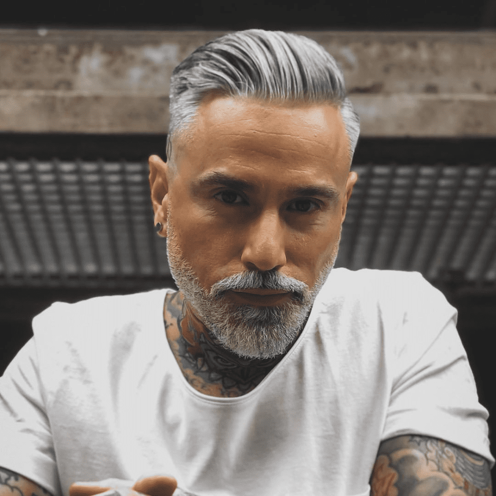 A mature man with silver hair and a well-groomed beard showcases a sleek pompadour slick back haircut. His hair is meticulously styled back, giving a polished and refined appearance. He is wearing a white t-shirt that highlights his muscular build and tattoos visible on his neck and arms. The background features an industrial setting with a metal grating, adding an edgy vibe to his sophisticated look. His intense gaze and confident posture reflect his stylish and bold personality.