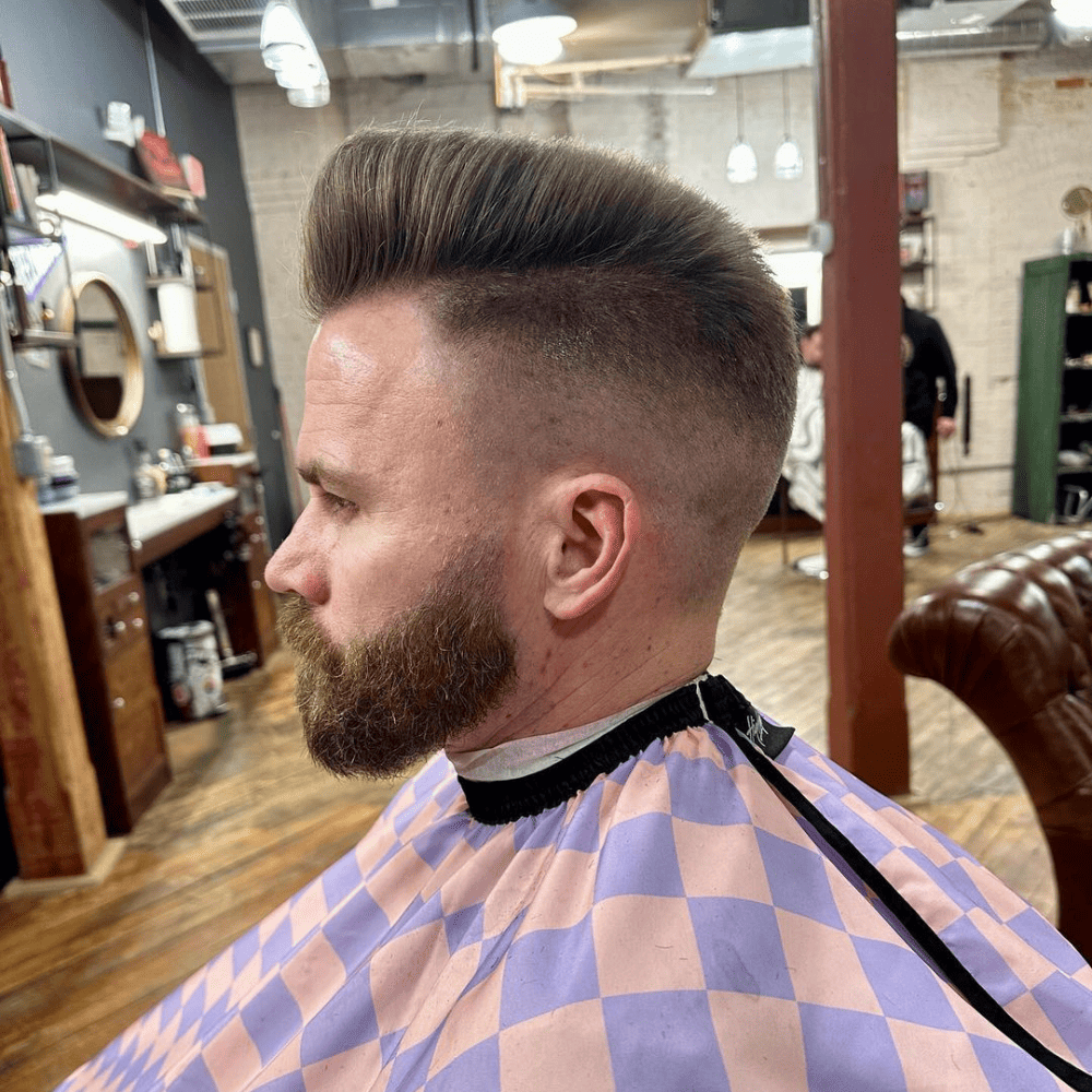 A man is seated in a barbershop, sporting a stylish quiff pompadour haircut. The sides of his head are shaved in a clean fade, while the top hair is voluminous and neatly styled upwards, creating a striking contrast. His beard is well-groomed, complementing the sophisticated look of his haircut. The barbershop has a rustic ambiance with wooden flooring and modern equipment, enhancing the overall aesthetic. The man’s expression reflects satisfaction with his fresh, fashionable haircut.